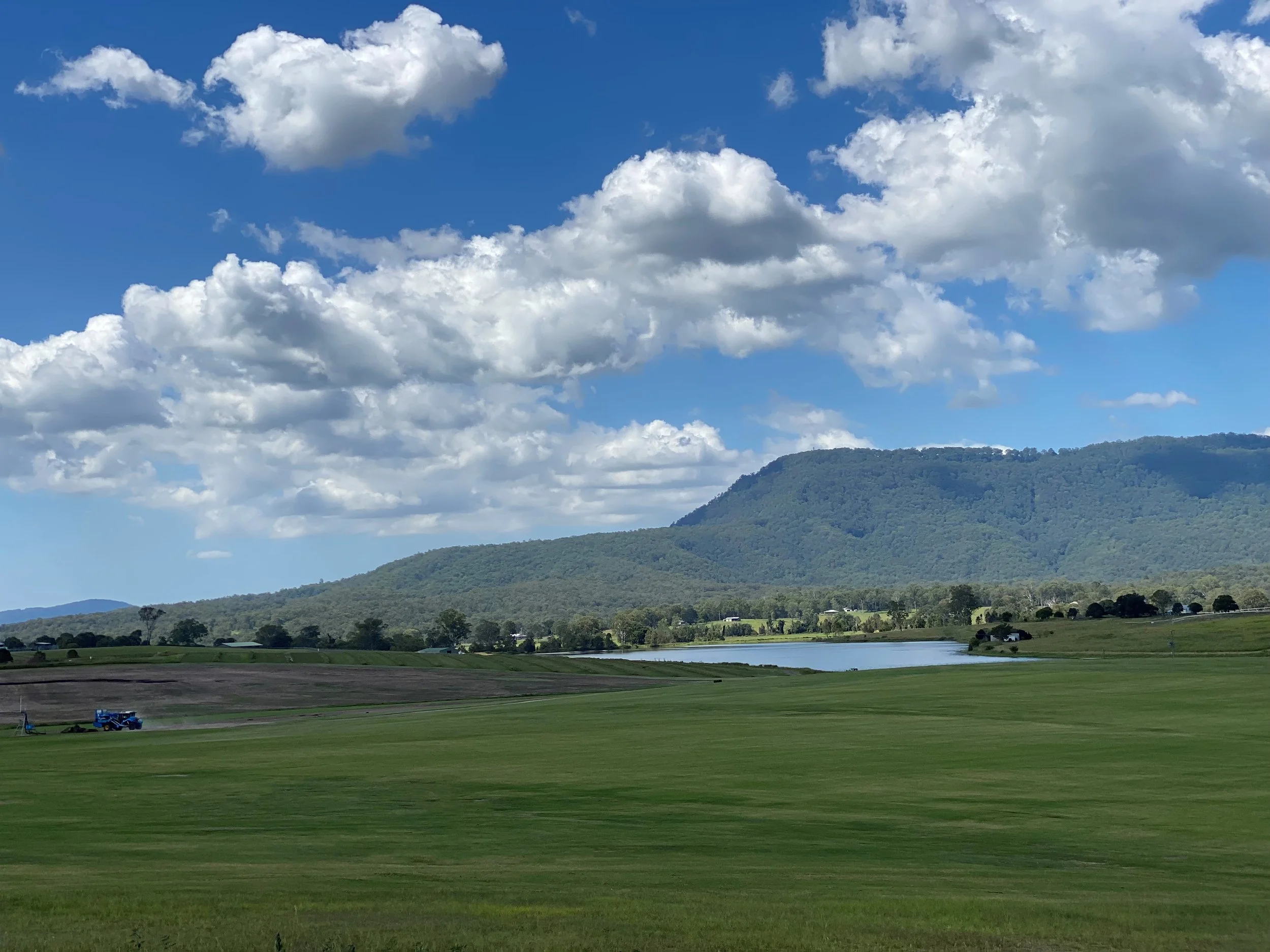 A scenic landscape featuring a blue sky with scattered white clouds, a green forested mountain, a lake, and a clear grassy area with some machinery and small buildings in the foreground.