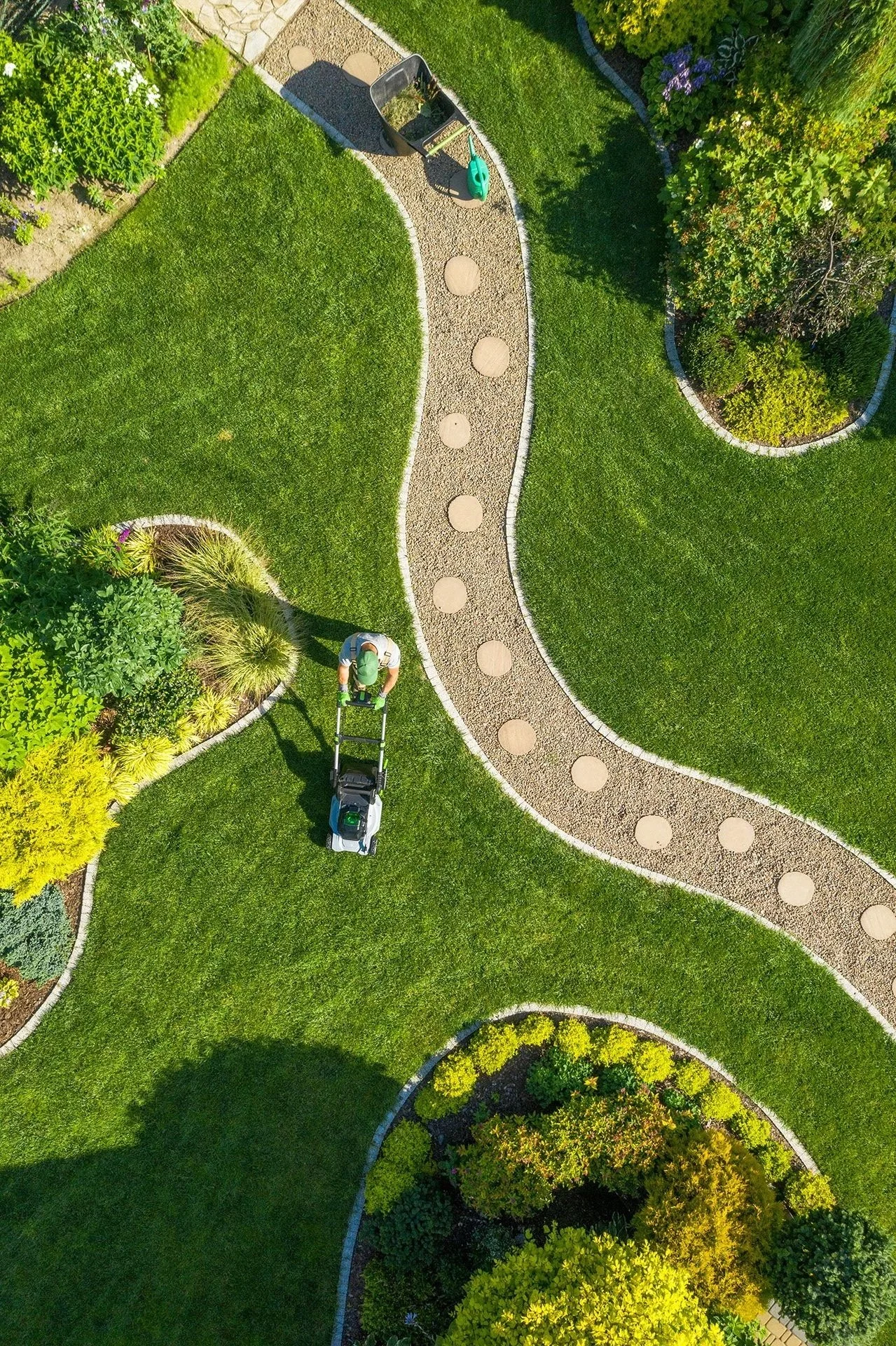 An overhead view of a well-maintained garden with a winding gravel pathway, green grass, and various colorful plants and bushes. A person is mowing the lawn near a small garden bed.