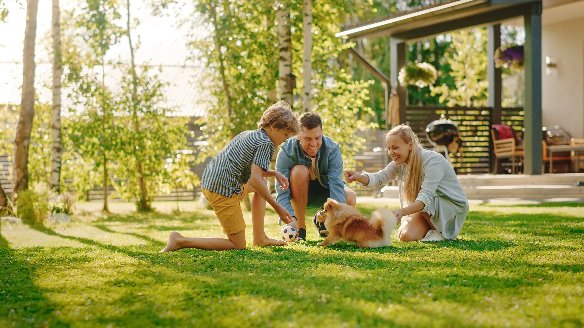 Family of three and a dog playing with a soccer ball on the grass in a sunny backyard, surrounded by trees and a house in the background.