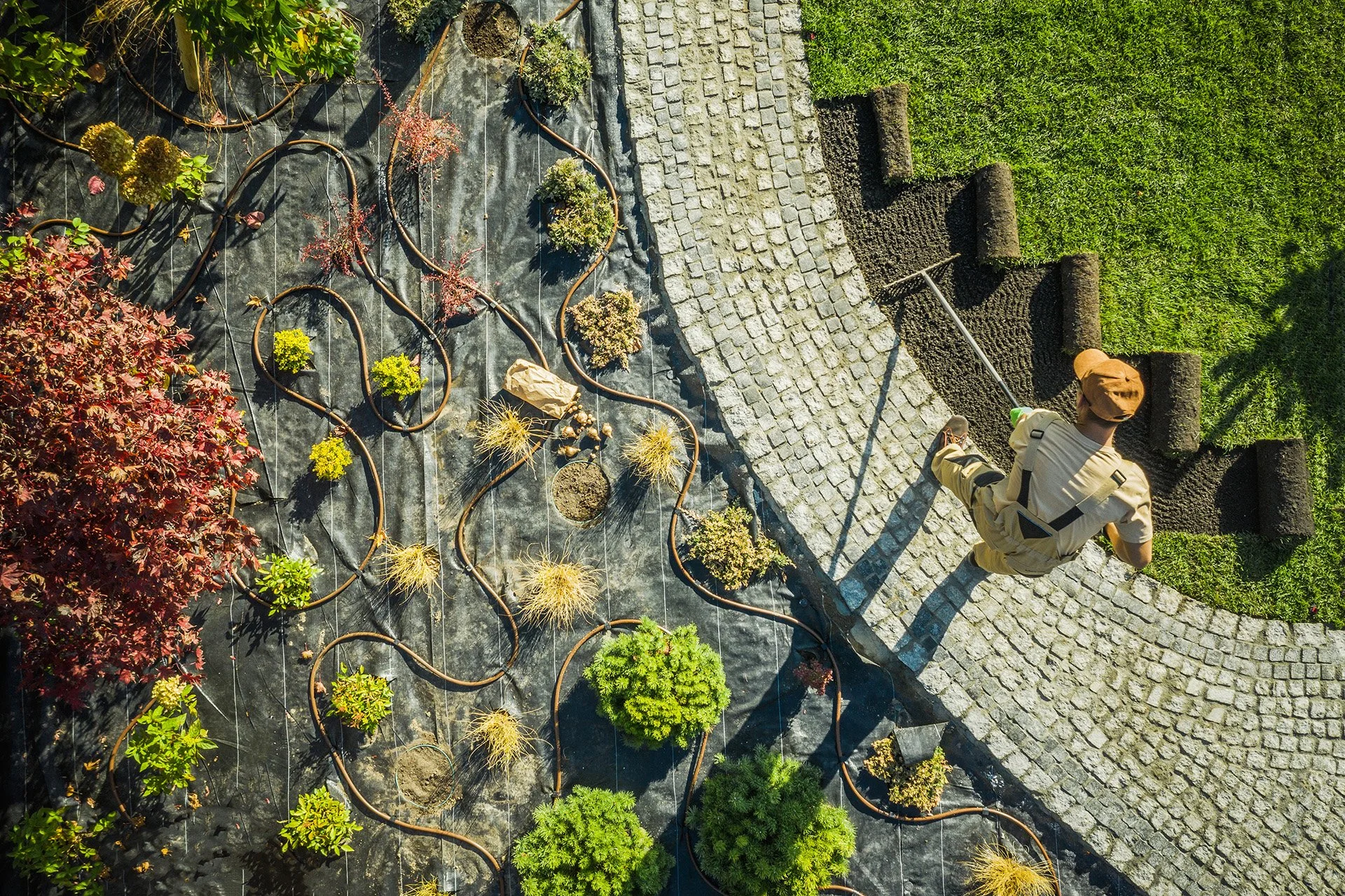 An overhead view of a person gardening on a paved pathway, with rows of plants in pots lined along the black ground cover and a gravel section nearby. The person is wearing a hat and light-colored clothing, using a gardening tool to tend to the soil.