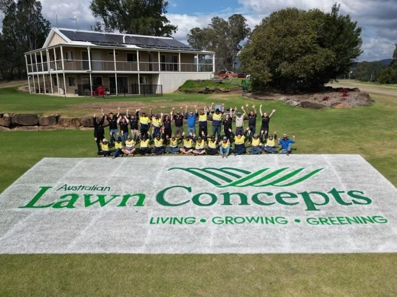 Group of people posing on a lawn in front of a large grass logo for Australian Lawn Concepts. The logo reads 'Living, Growing, Greenings' and is painted on the grass. A house and trees are in the background.