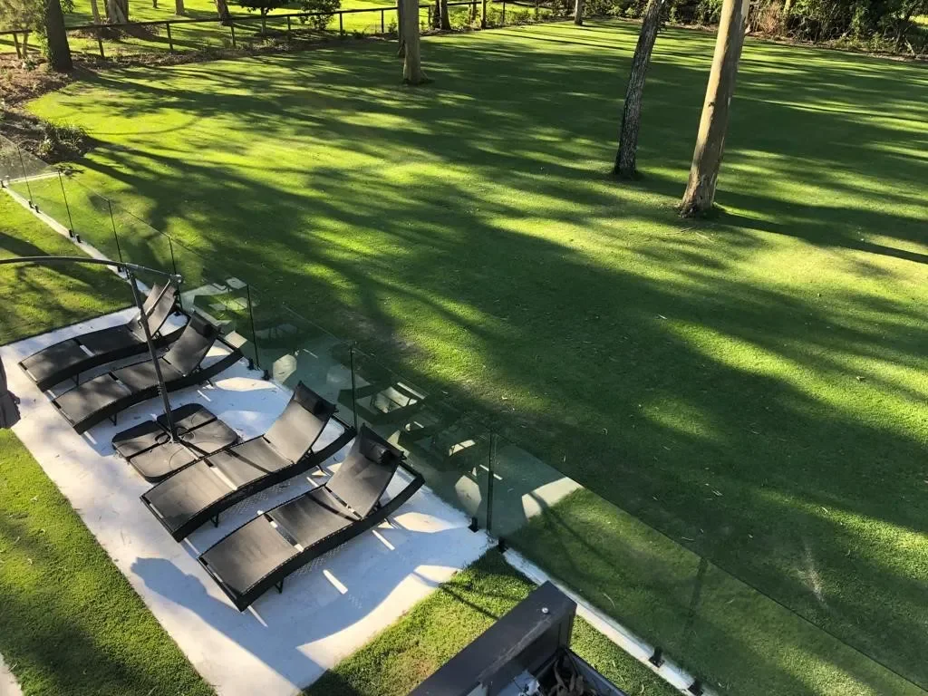 Outdoor patio with black lounge chairs and glass railing overlooking a grassy backyard with trees casting shadows.