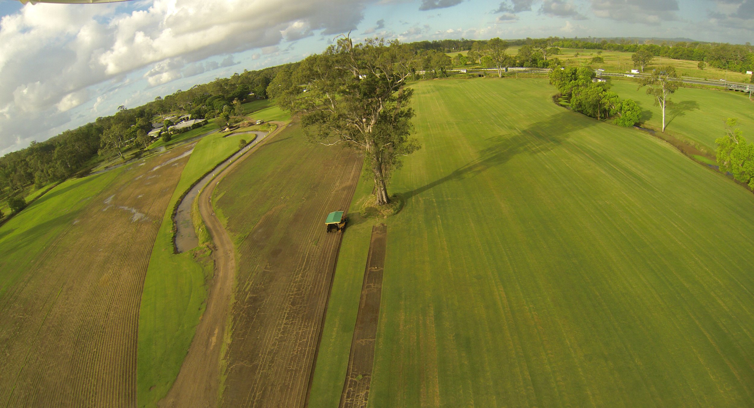 Aerial view of Glenview Turf farm with large trees, dams, and a dirt road. There are fields in various stages of cultivation and a tractor working on one of the fields.
