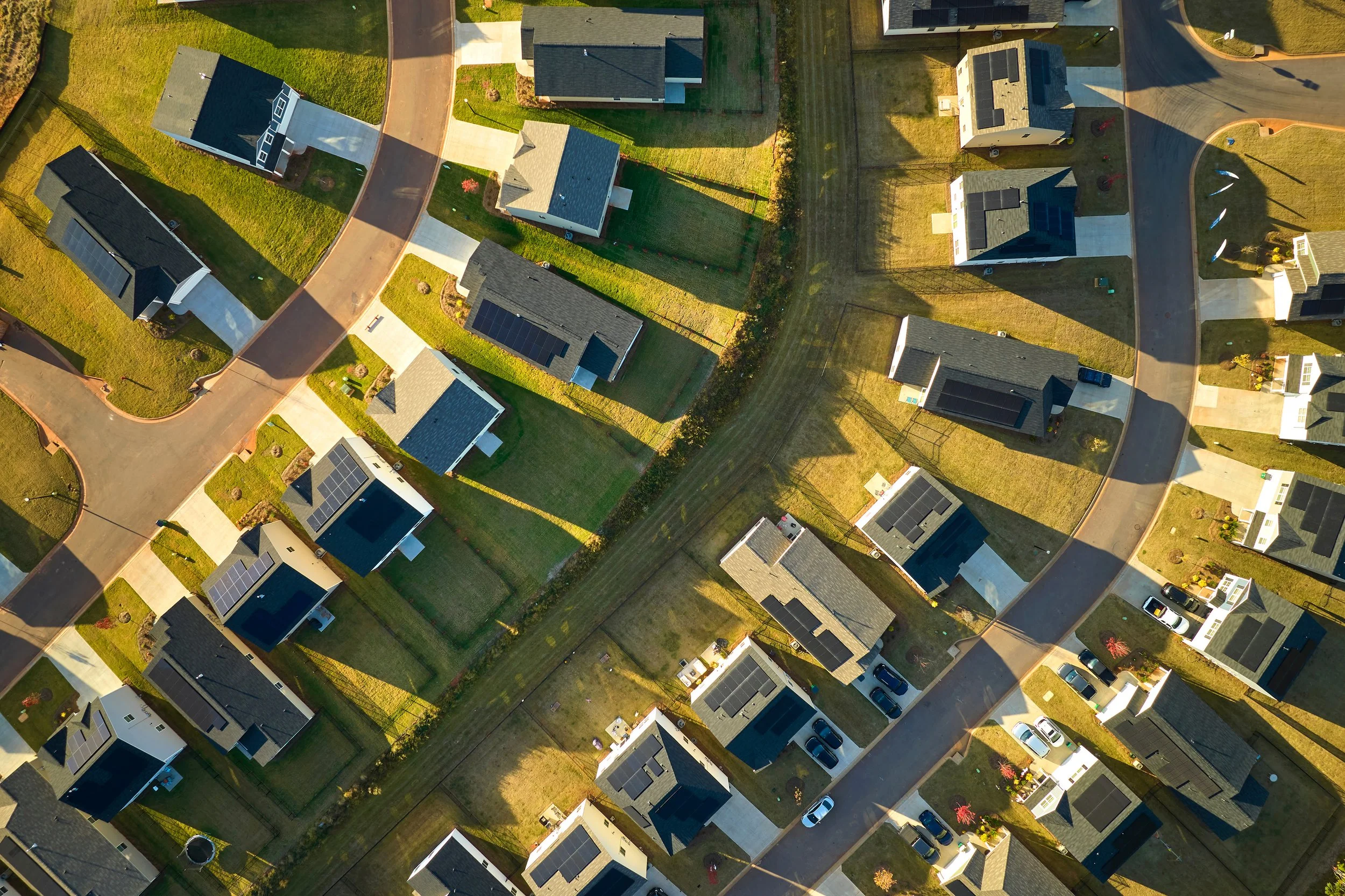 An aerial view of a neighborhood showing houses with solar panels, fenced yards, and streets with parked cars.