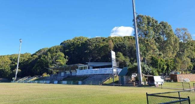 View of a sports stadium on a sunny day with a grassy field, tiered seating, tall light poles, and trees in the background.
