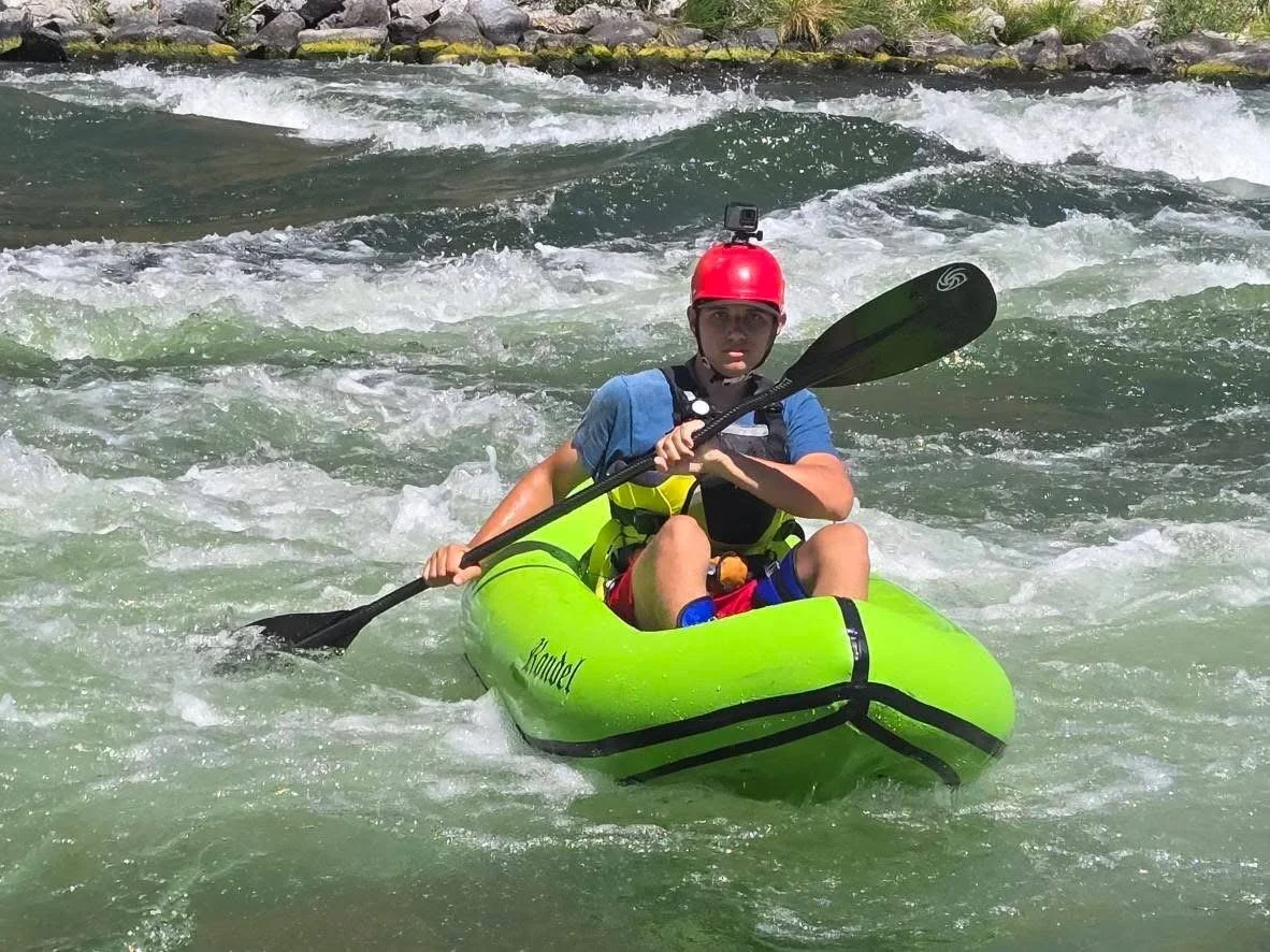 A person kayaking in white water rapids, wearing a red helmet with a GoPro camera, blue shirt, life jacket, and holding a black paddle in a green kayak.