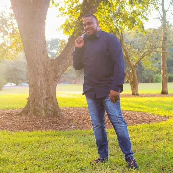 A man standing in a park near a large tree, smiling and holding a phone to his ear, wearing a dark blue shirt and jeans, during daylight.