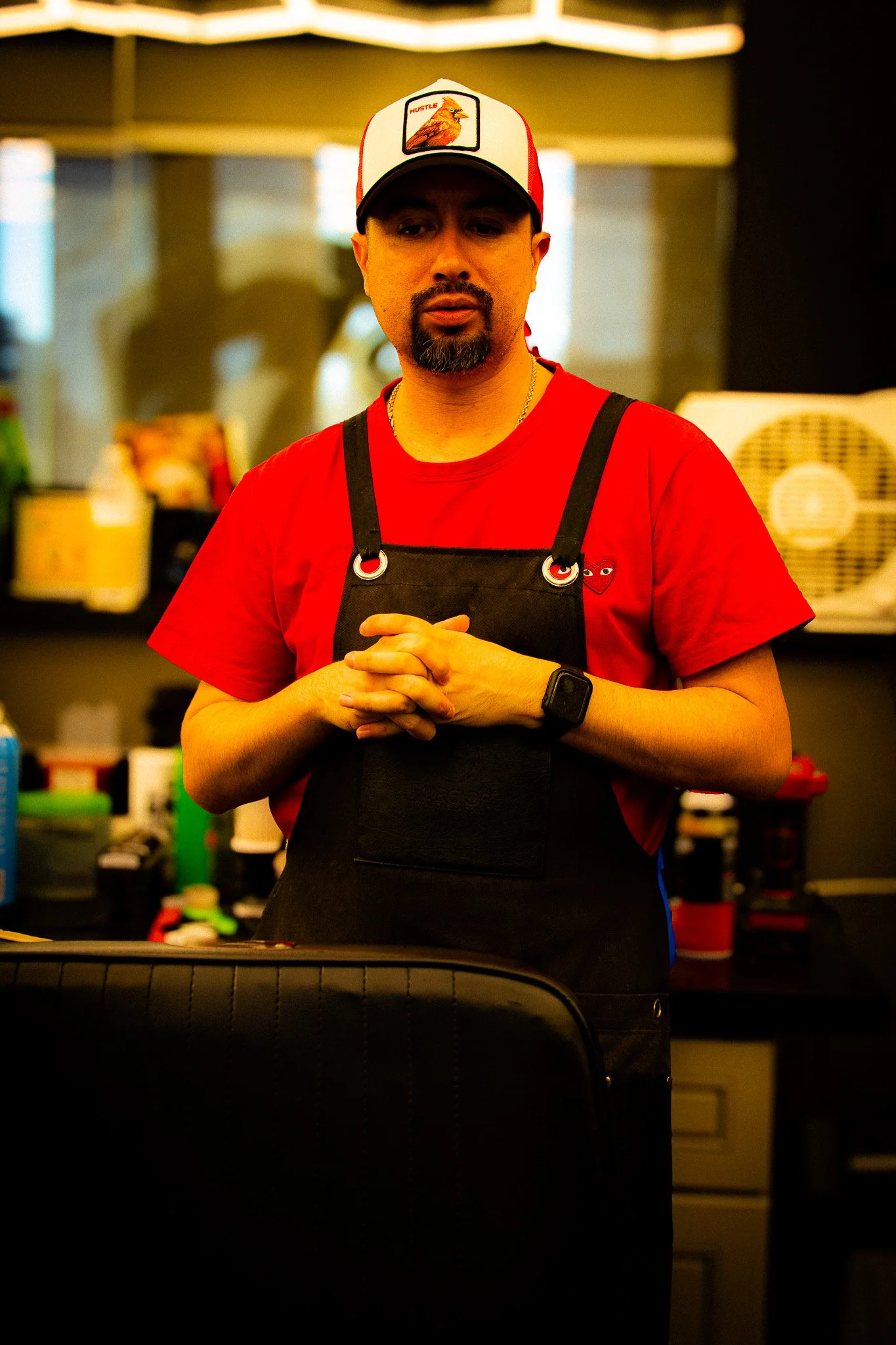 A man with a beard and mustache wearing a red t-shirt, black apron, and a white trucker cap with a bird image, standing in a kitchen or workspace with various supplies in the background.