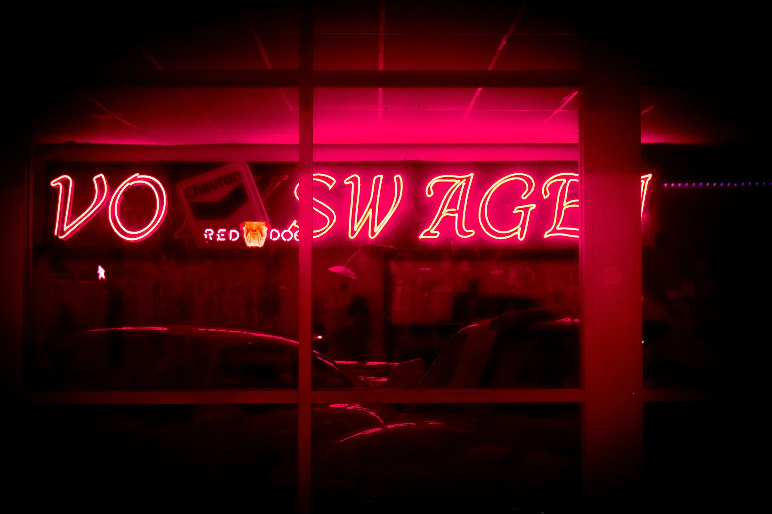 A neon sign in pink reads 'VO SWAGE' with partially visible words and a Chevron logo in the background, viewed through a glass window at night.