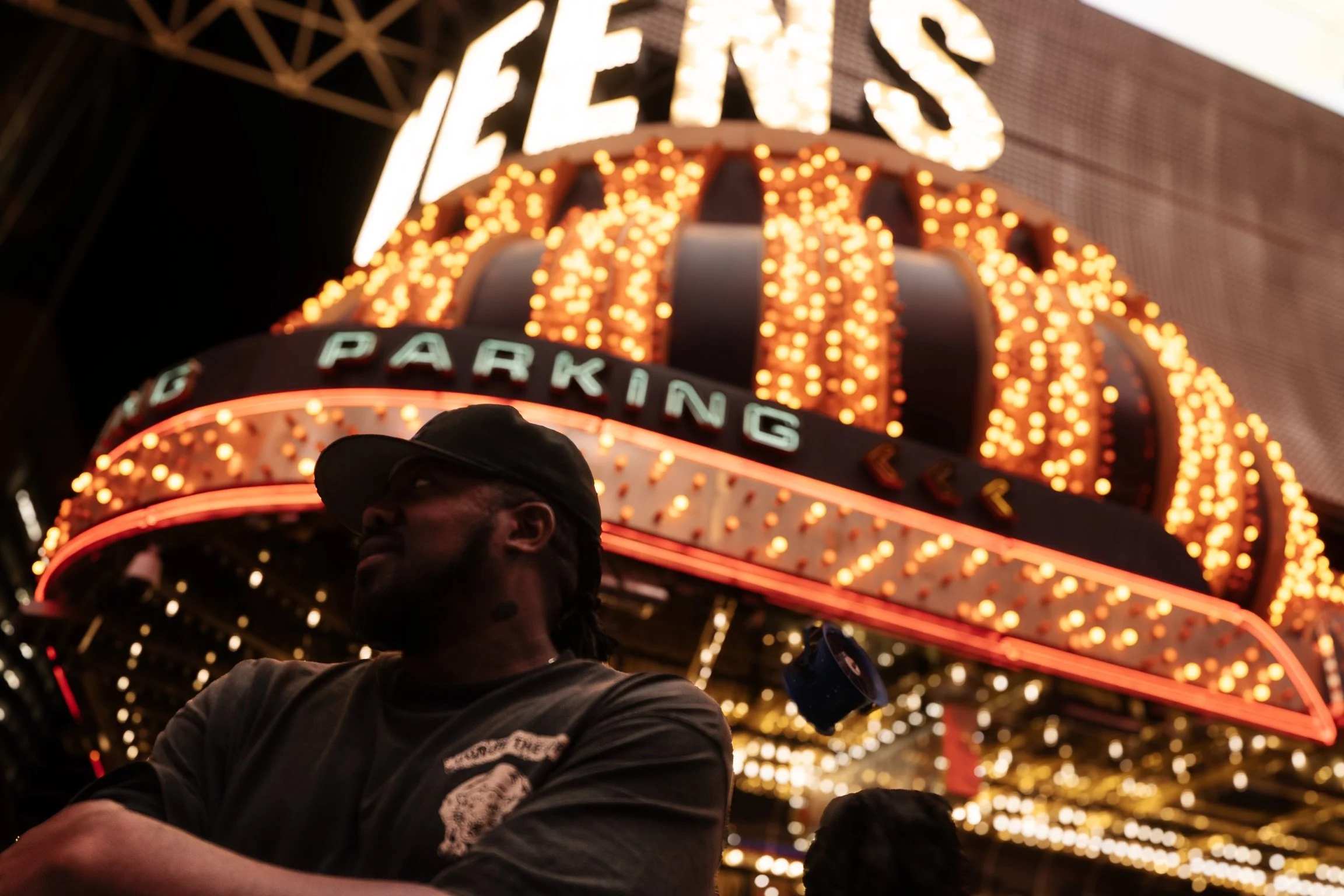 A man wearing a baseball cap and dark shirt stands in front of a brightly lit amusement park or arcade with a sign that reads 'GAMES' and 'PARKING.' The background is filled with warm, glowing lights.