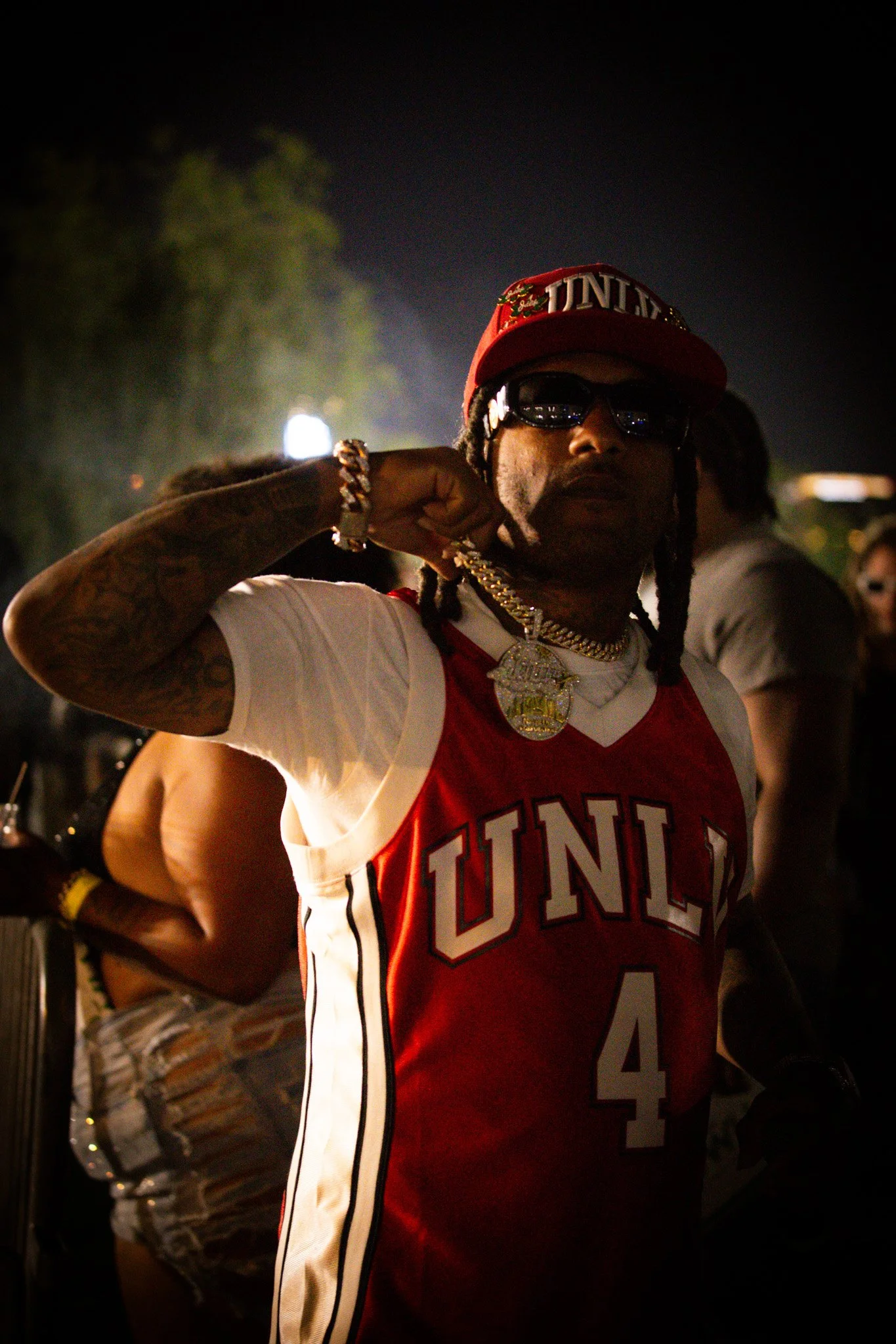 A man with long dreadlocks wearing sunglasses, a red cap, and a red and white basketball jersey displaying 'UNLV' and the number 4, holding a gold chain with a large pendant.