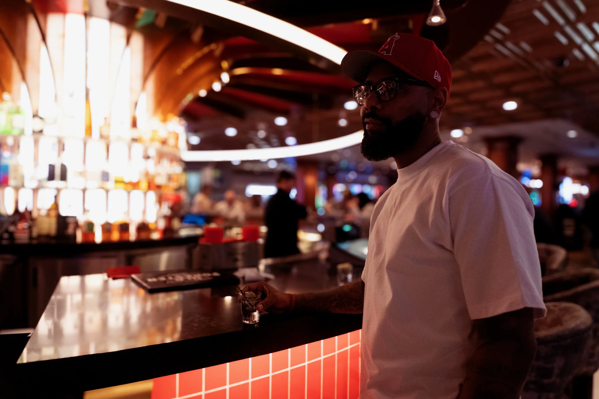 A man with a beard, wearing a red cap, glasses, a white t-shirt, and a wristband, standing at a bar in a dimly lit nightclub or bar with a modern, stylish interior, holding a glass of drink.