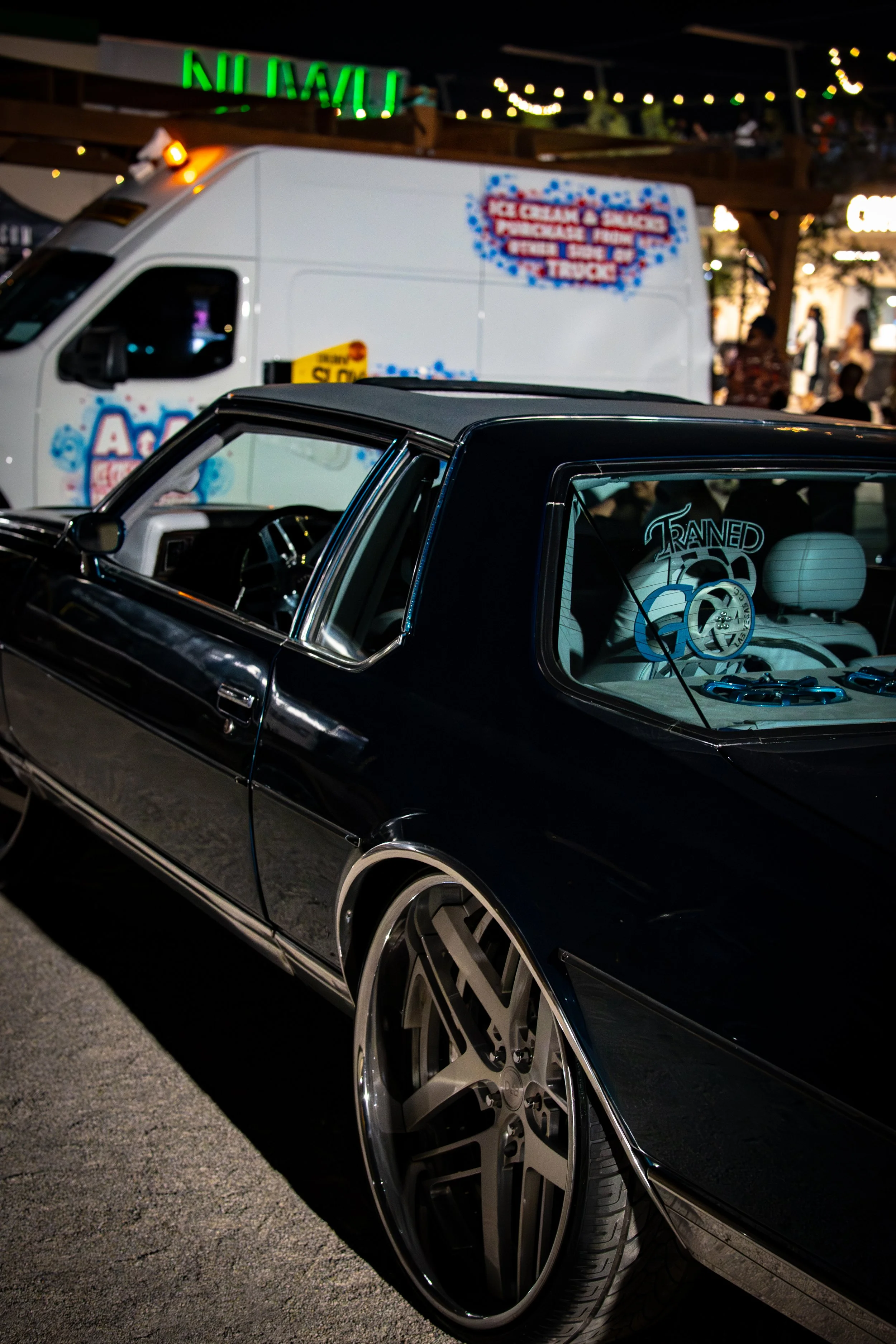 A black vintage car with oversized rims parked at a night event with a white ice cream truck and string lights in the background.