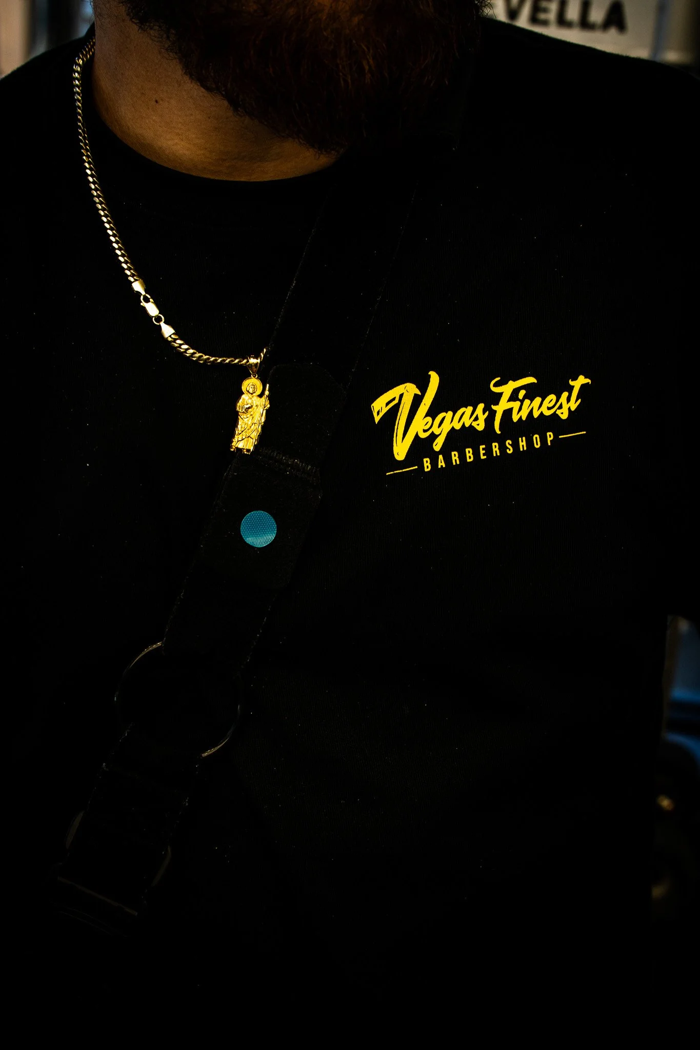 Close-up of a person wearing a black shirt with yellow logo reading 'Vegas Finest Barbershop,' a gold necklace with a religious pendant, and a shoulder strap.