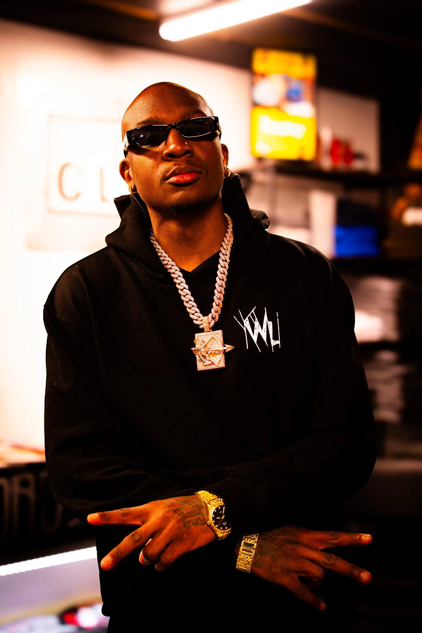 Man wearing black hoodie, sunglasses, and gold jewelry, including a chain with a pendant, a watch, and a bracelet, standing indoors with blurred background.