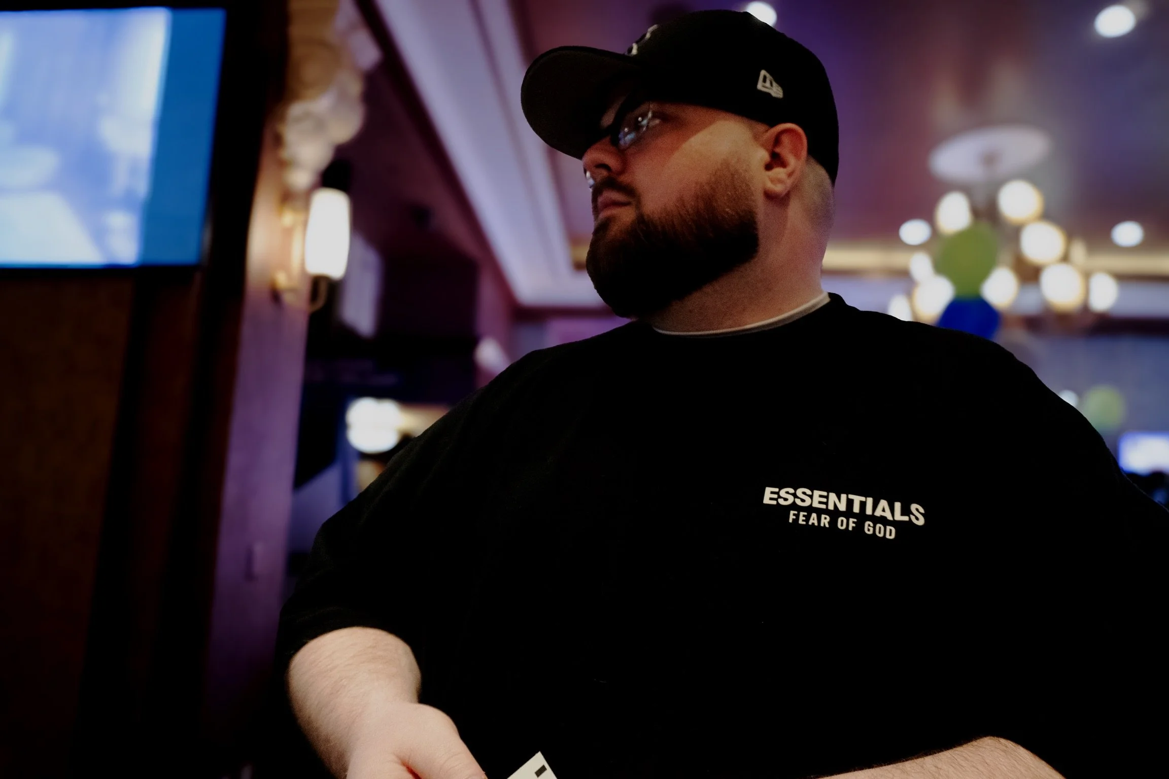 A man with glasses, beard, and black cap looking to the side in a dimly lit indoor setting, wearing a black shirt with white text that reads "ESSENTIALS FEAR OF GOD."