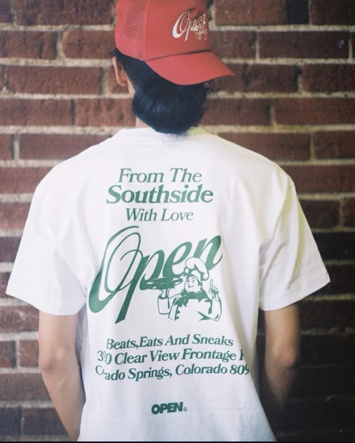 Back of a person wearing a white T-shirt with green text and a red baseball cap with white text, standing in front of a brick wall.