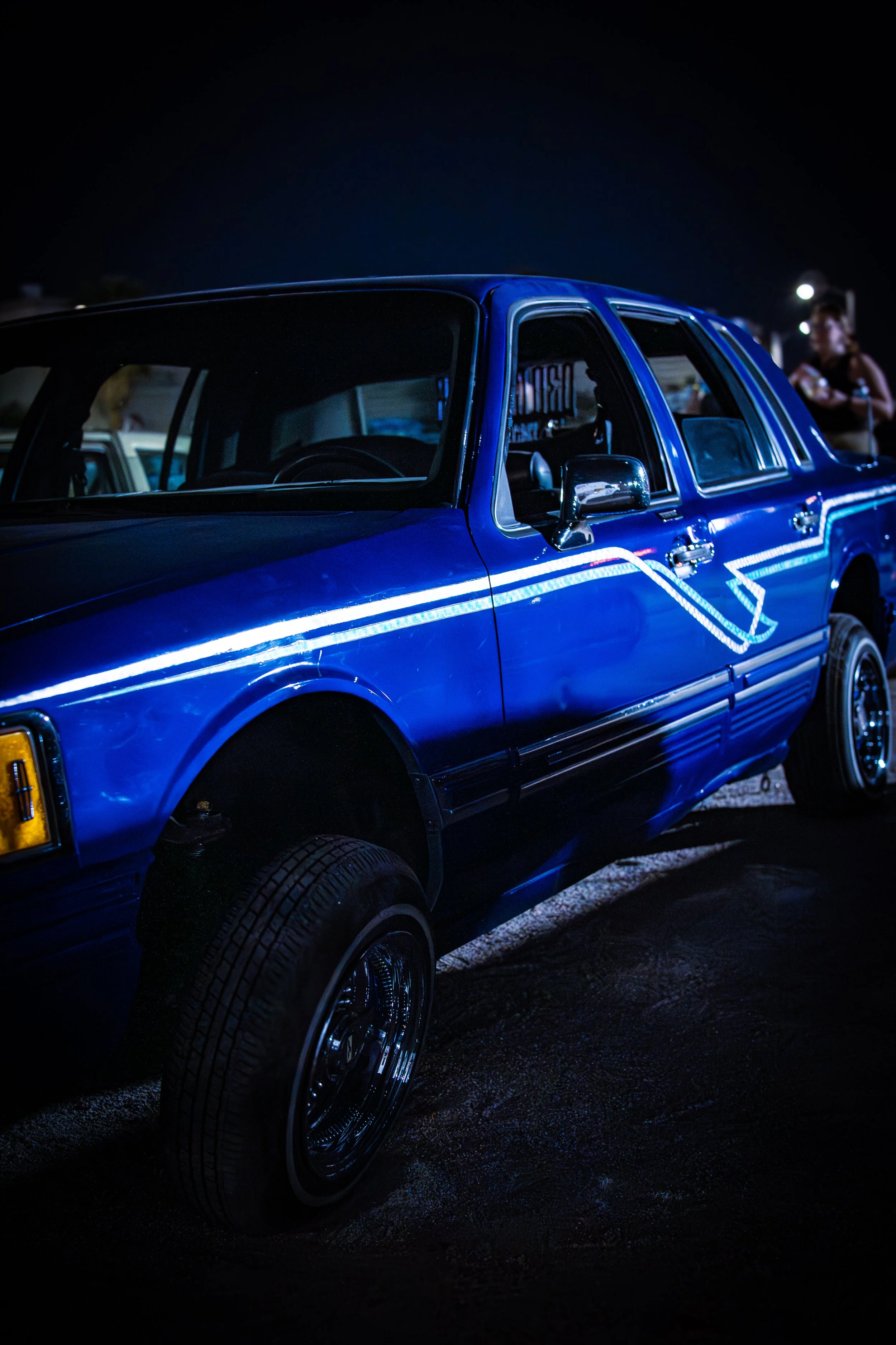 A blue vintage car parked outdoors at night, with reflections along its body, and a woman standing in the background.