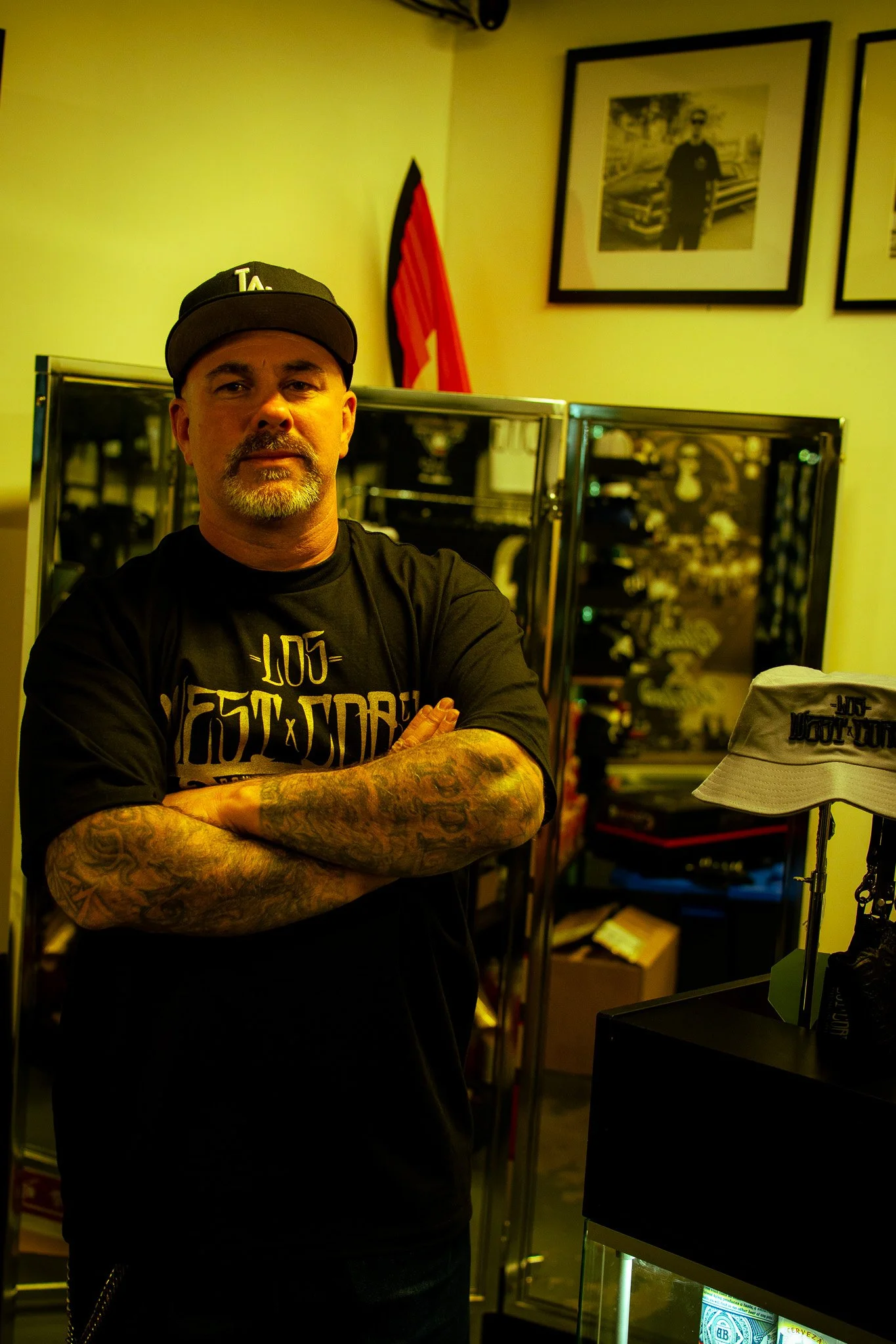 A man with tattoos on his arms standing with arms crossed in a store. He's wearing a black cap and black t-shirt with yellow and white designs. Behind him are display cabinets with items and a framed black-and-white photo on the wall.