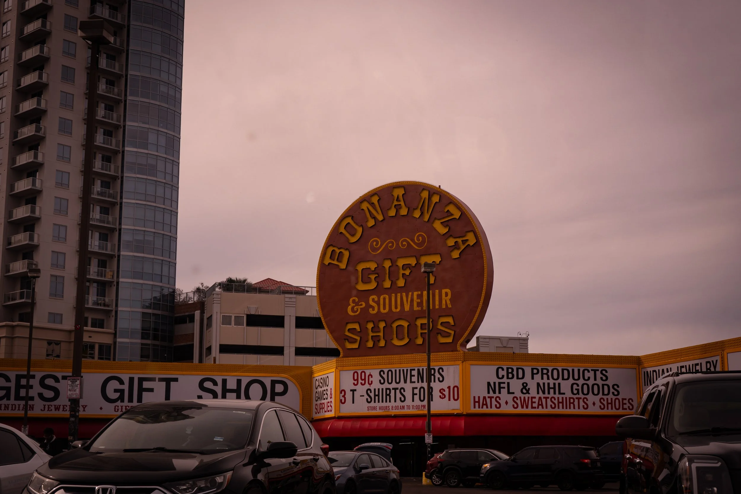 Large sign for Bonanza Gift & Souvenir Shops with cars parked in front. In the background, tall buildings and a cloudy sky.