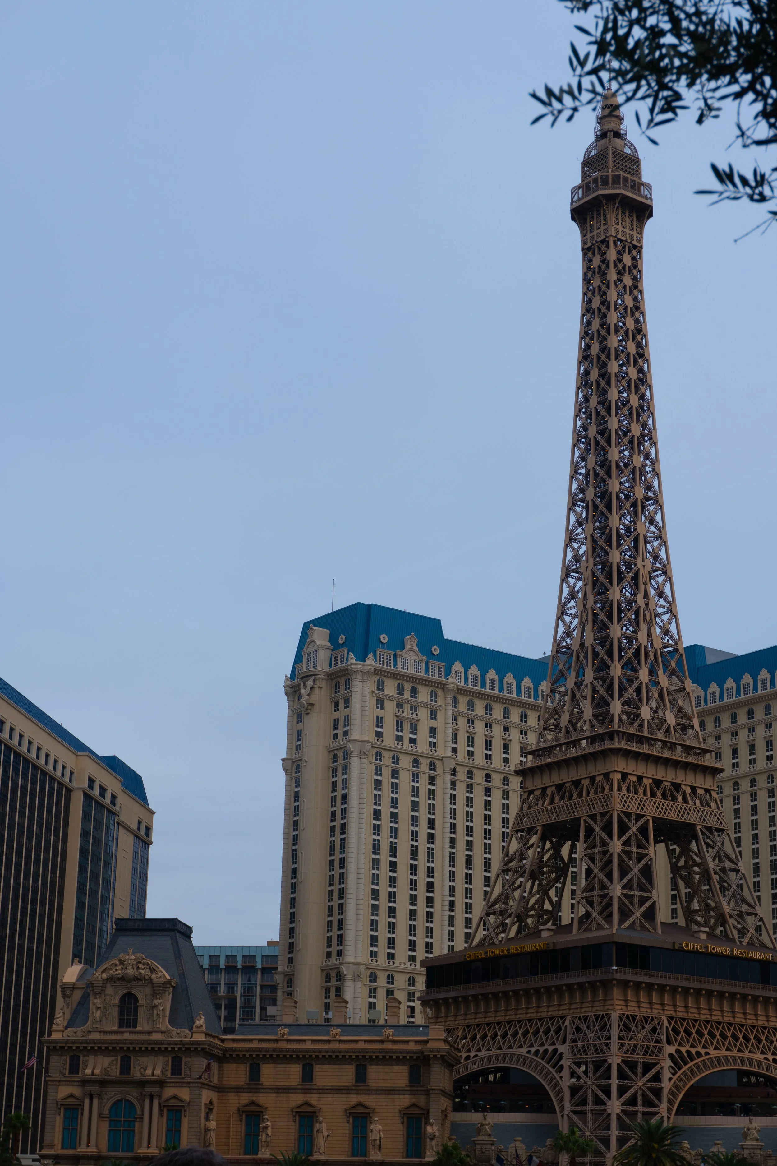 Replica of the Eiffel Tower at a Las Vegas hotel with surrounding buildings, including a hotel with a blue roof, and palm trees in front.