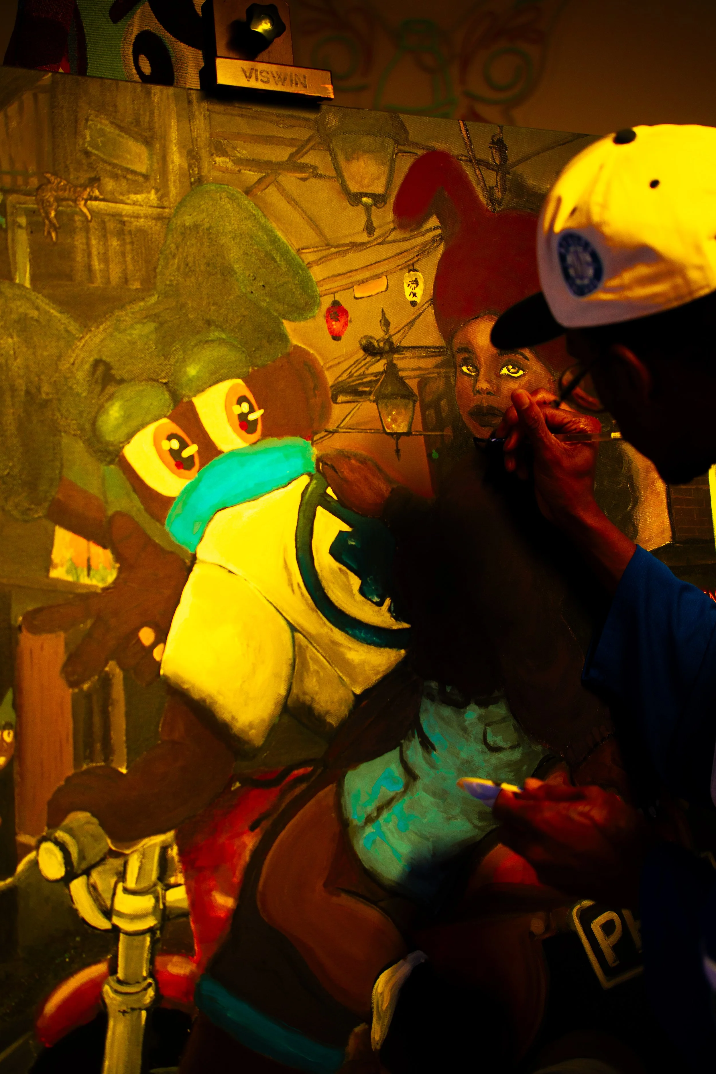 An artist is painting a colorful mural featuring a cartoonish character with big eyes and a blue face mask, alongside a realistic portrait of a woman with dark hair and expressive eyes, in a dimly lit indoor setting.