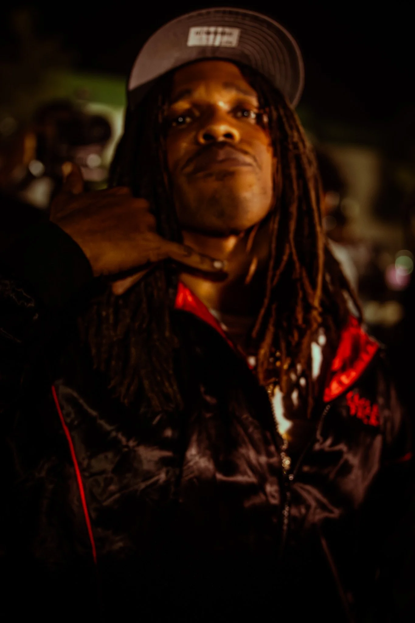 A man with dreadlocks wearing a baseball cap and a black jacket with red accents, making a casual hand gesture near his neck.