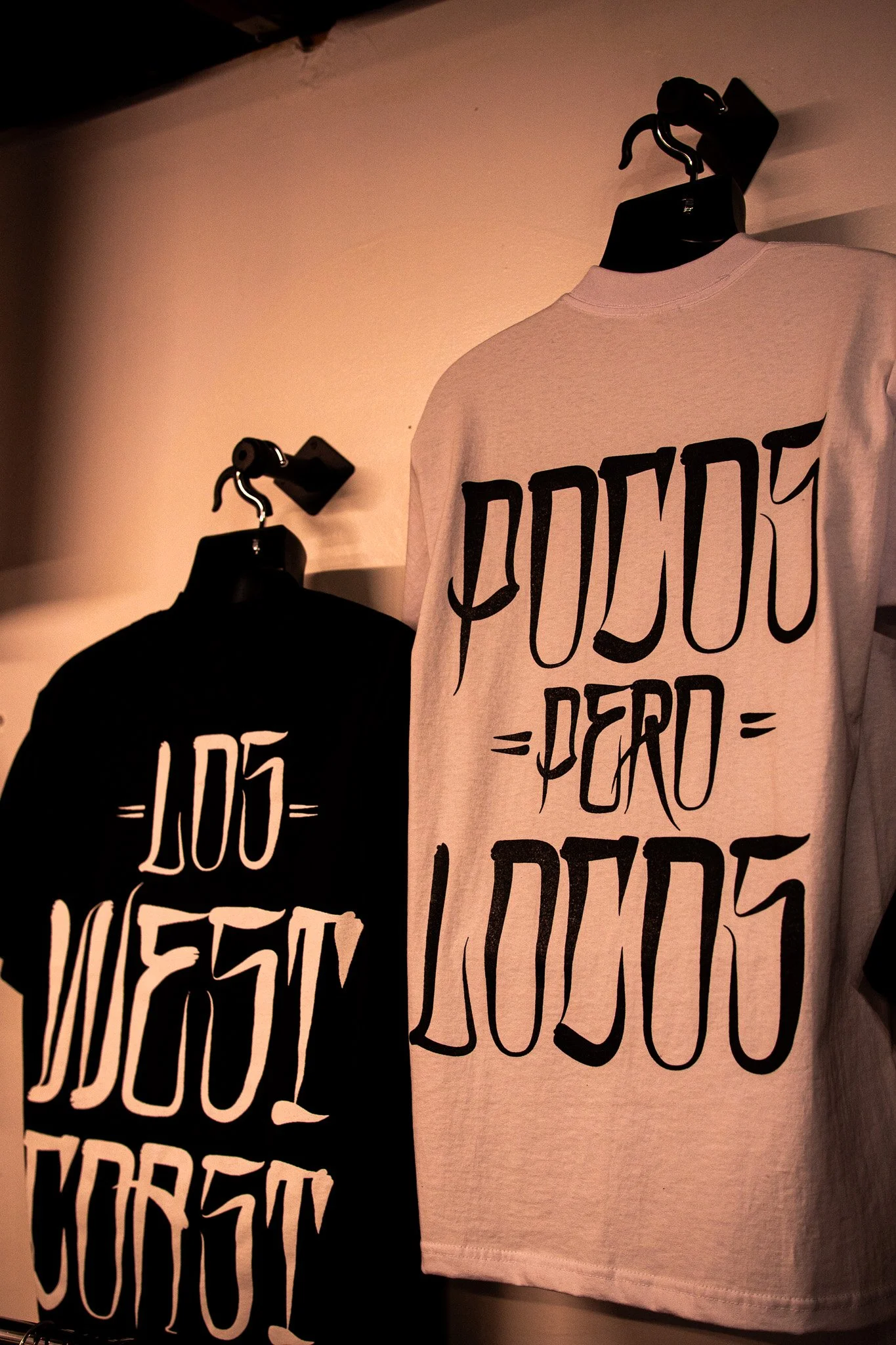 Two t-shirts hanging on black hangers on the wall, one black and one white, both with stylized black lettering.