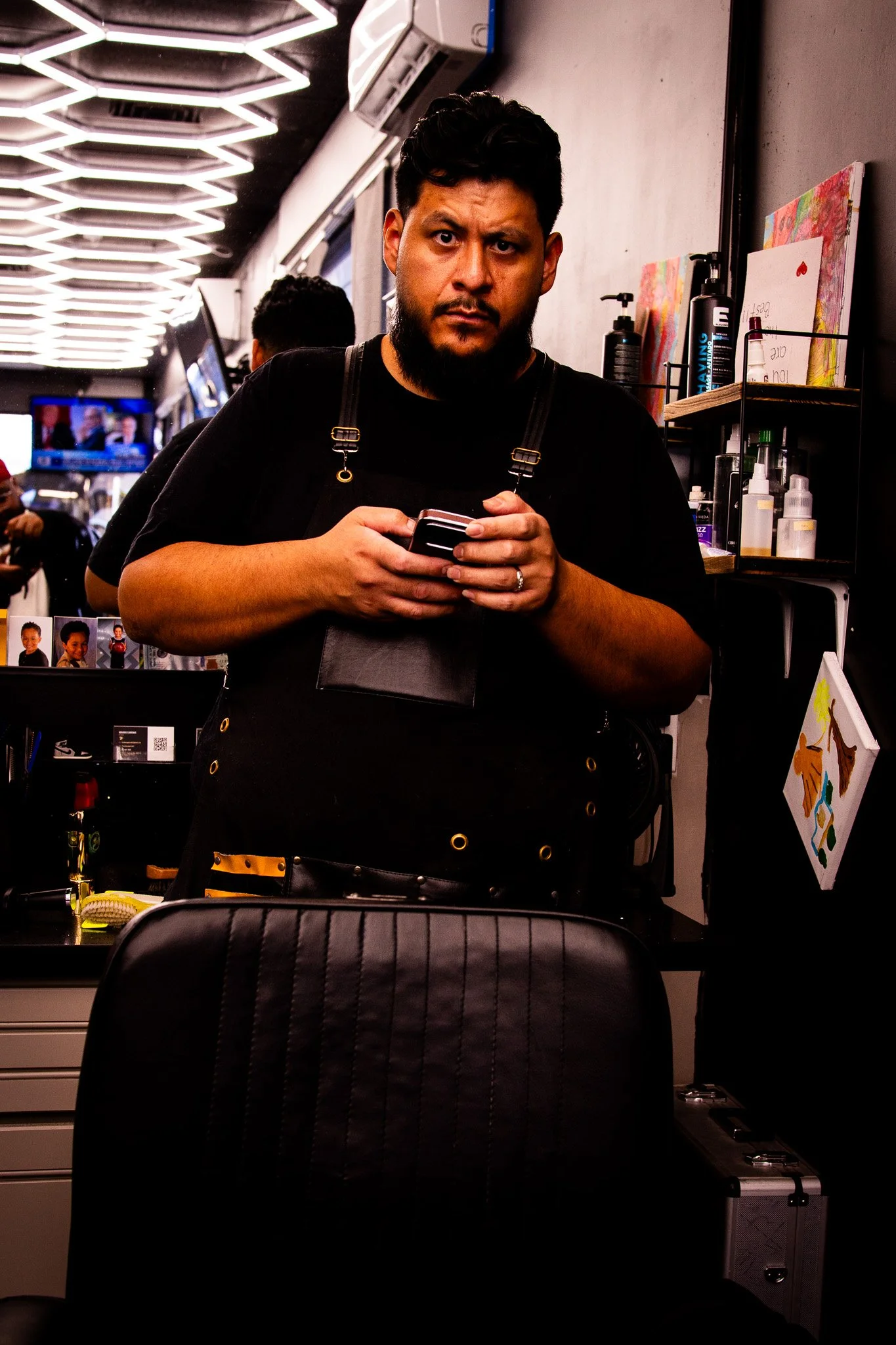 A man with a beard and black hair looking at the camera while holding a phone in a barbershop. The background shows bright ceiling lights, mirrors, and shelves with hair care products and artwork.