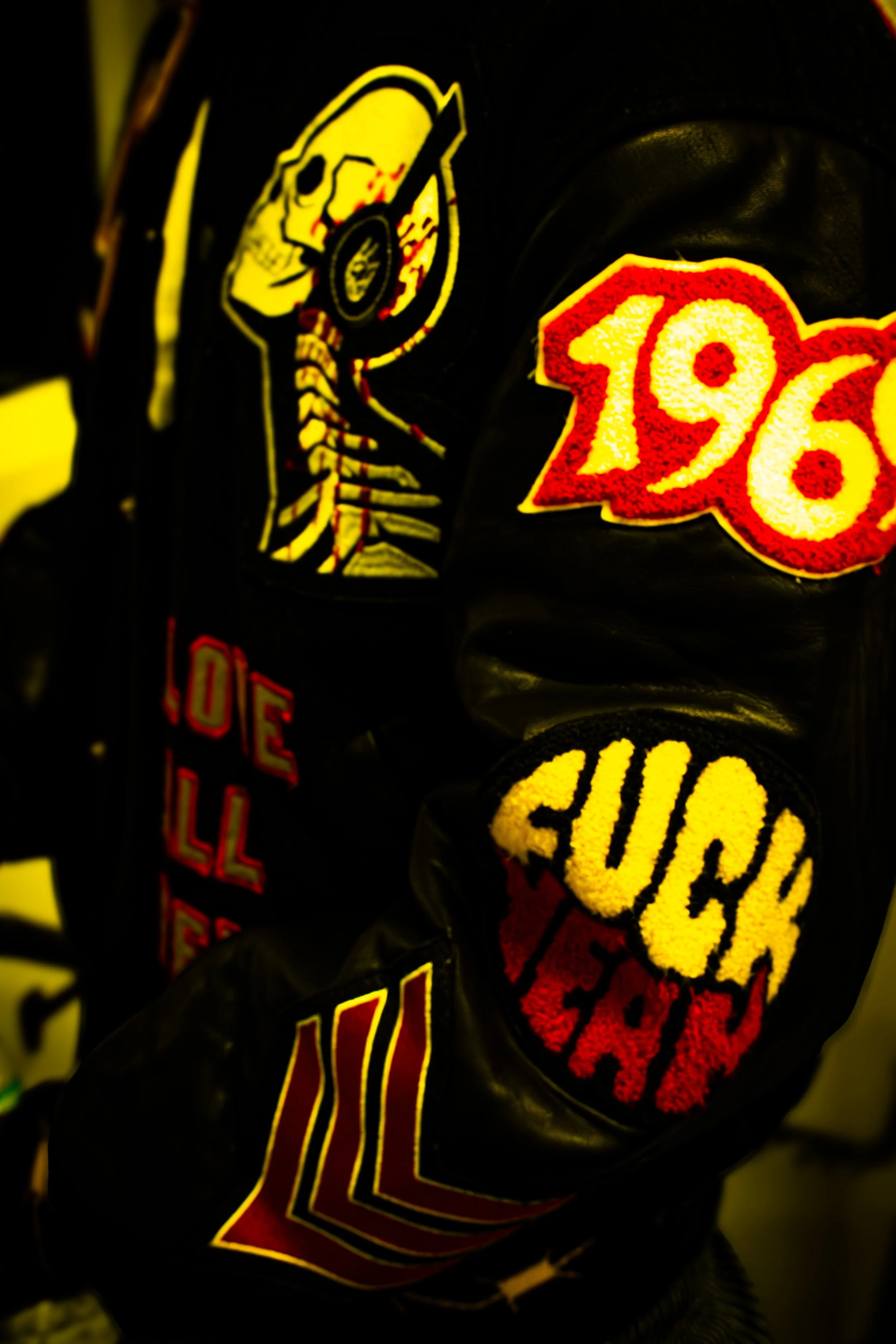 Close-up of a black leather jacket with patches. The patches include a skeleton wearing a helmet, a large red and yellow '1968' patch, a yellow and black 'QUICK' patch, and some red and yellow stripes.