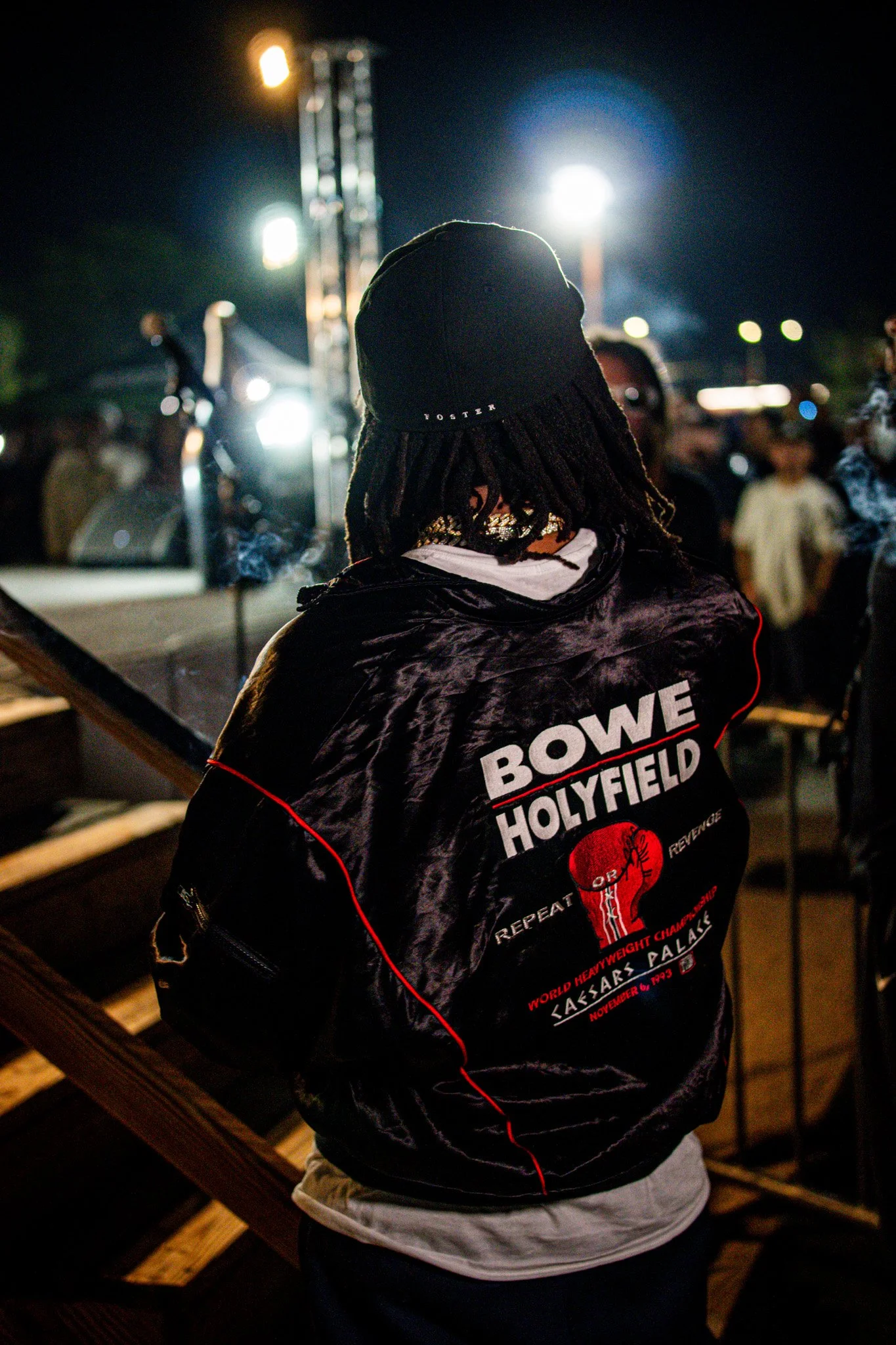 Person wearing a Bowery Holyfield jacket at an outdoor event at night with bright stage lights and people in the background.
