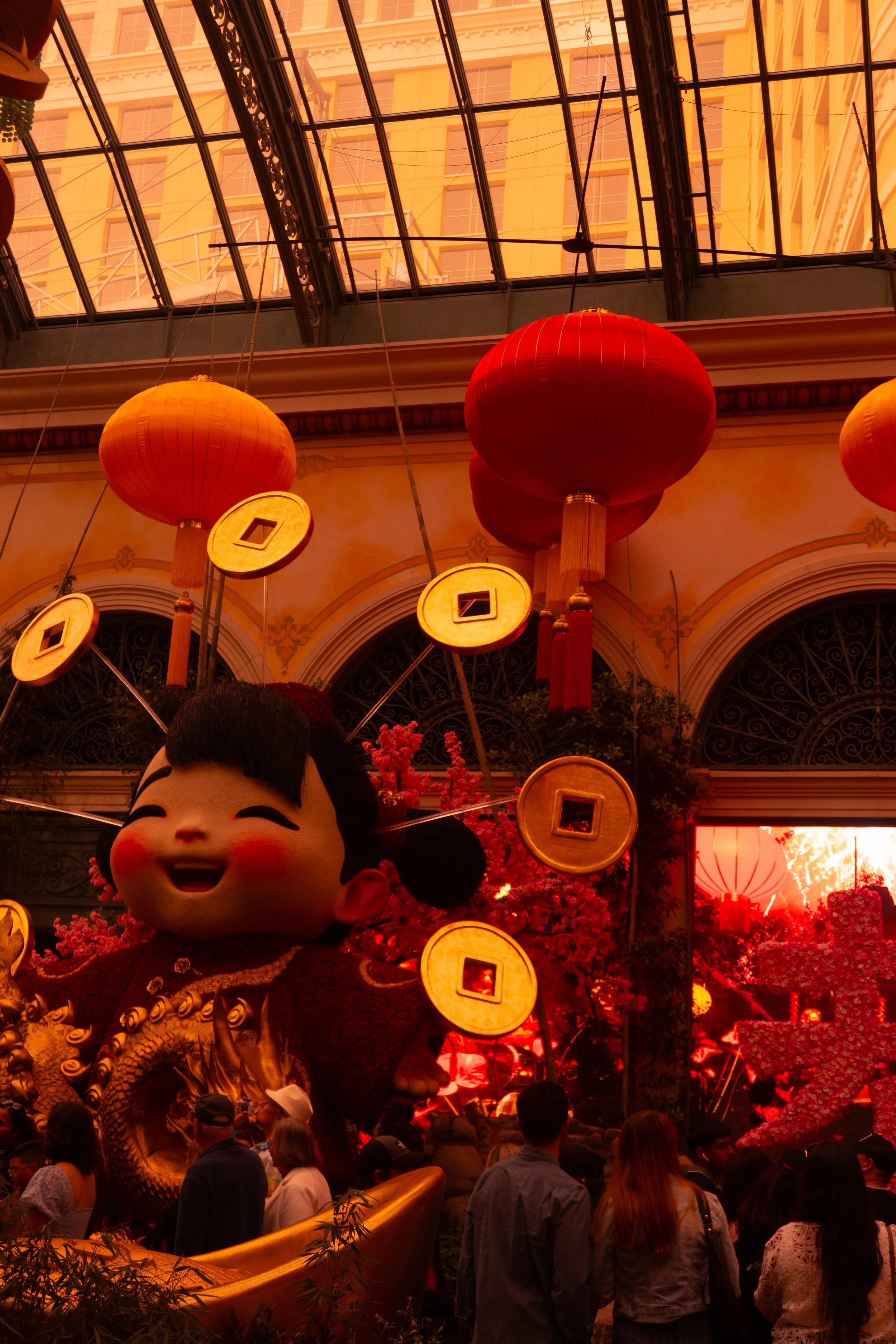 Indoor celebration with Chinese lanterns, a large smiling figure dressed in traditional Chinese attire, and decorative elements under a glass ceiling with a building visible outside.