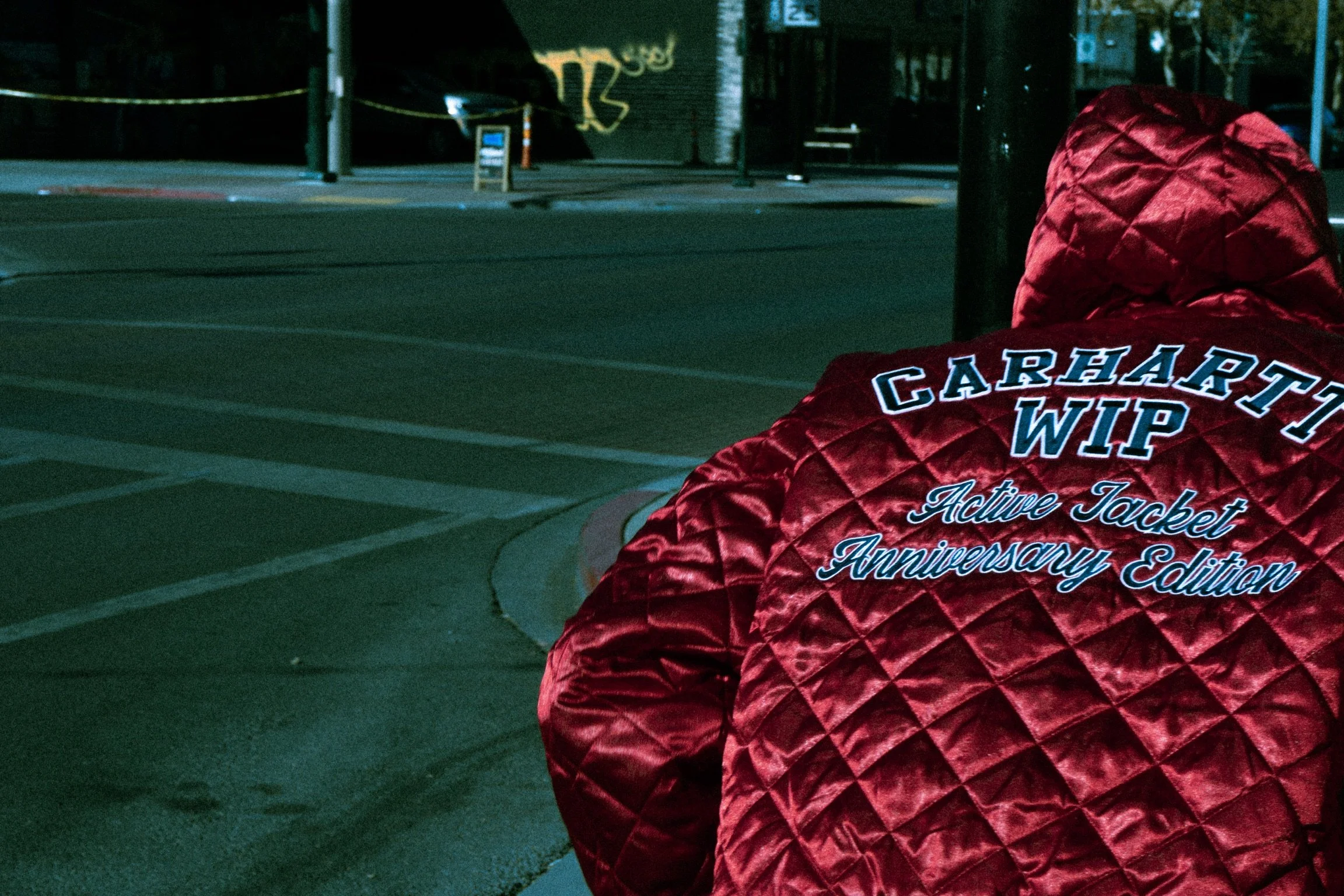 Person wearing a red quilted jacket with 'Carhartt WIP Active Jacket Anniversary Edition' embroidered on the back, sitting on a sidewalk at night, with a street scene in the background.