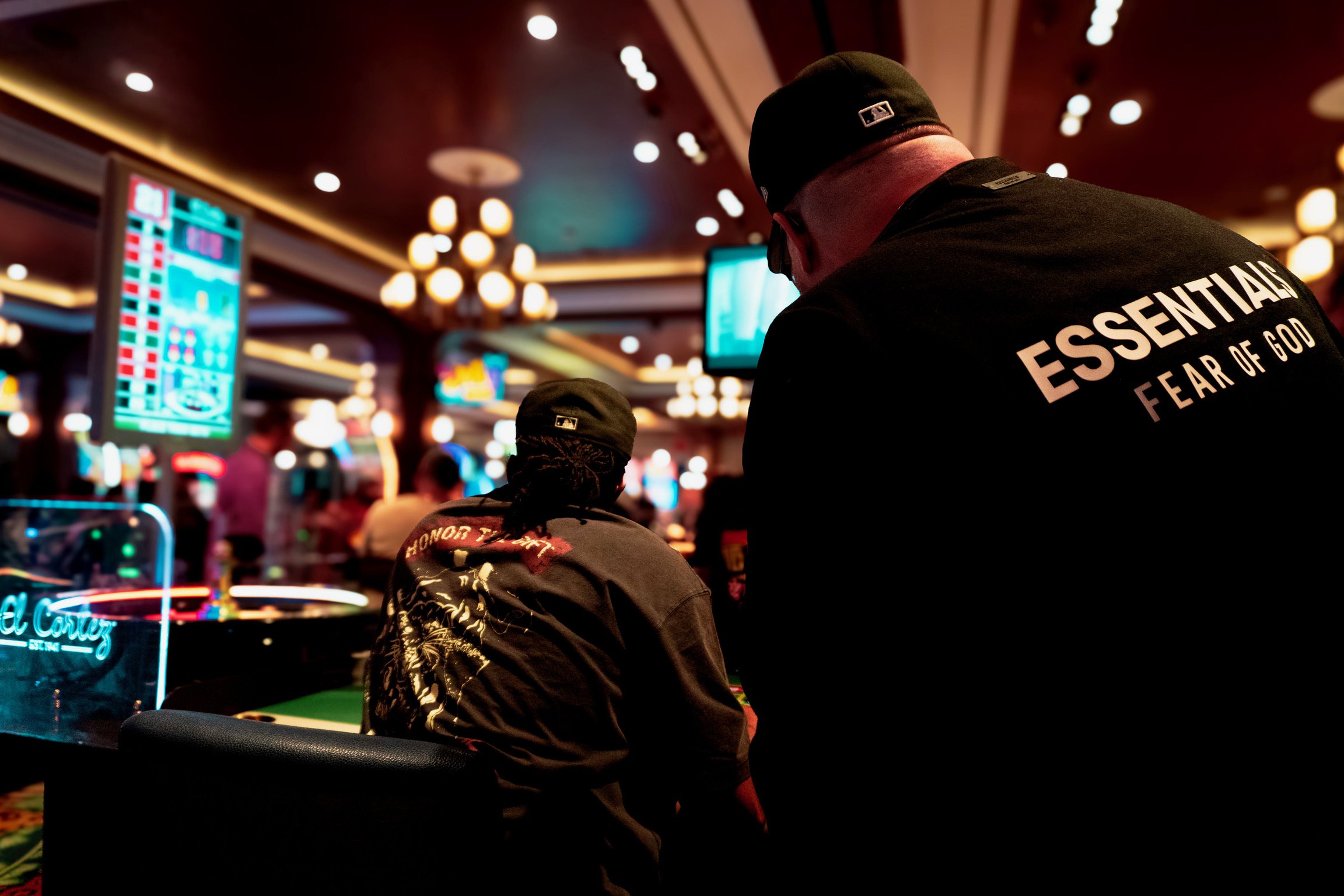 Two people sitting at a casino table in a dimly lit casino, with bright neon lights and screens in the background, one wearing a black cap and the other with a hoodie that reads "HONOR THE TRUE" and a black shirt with "ESSENTIAL FEAR OF GOD" printed 