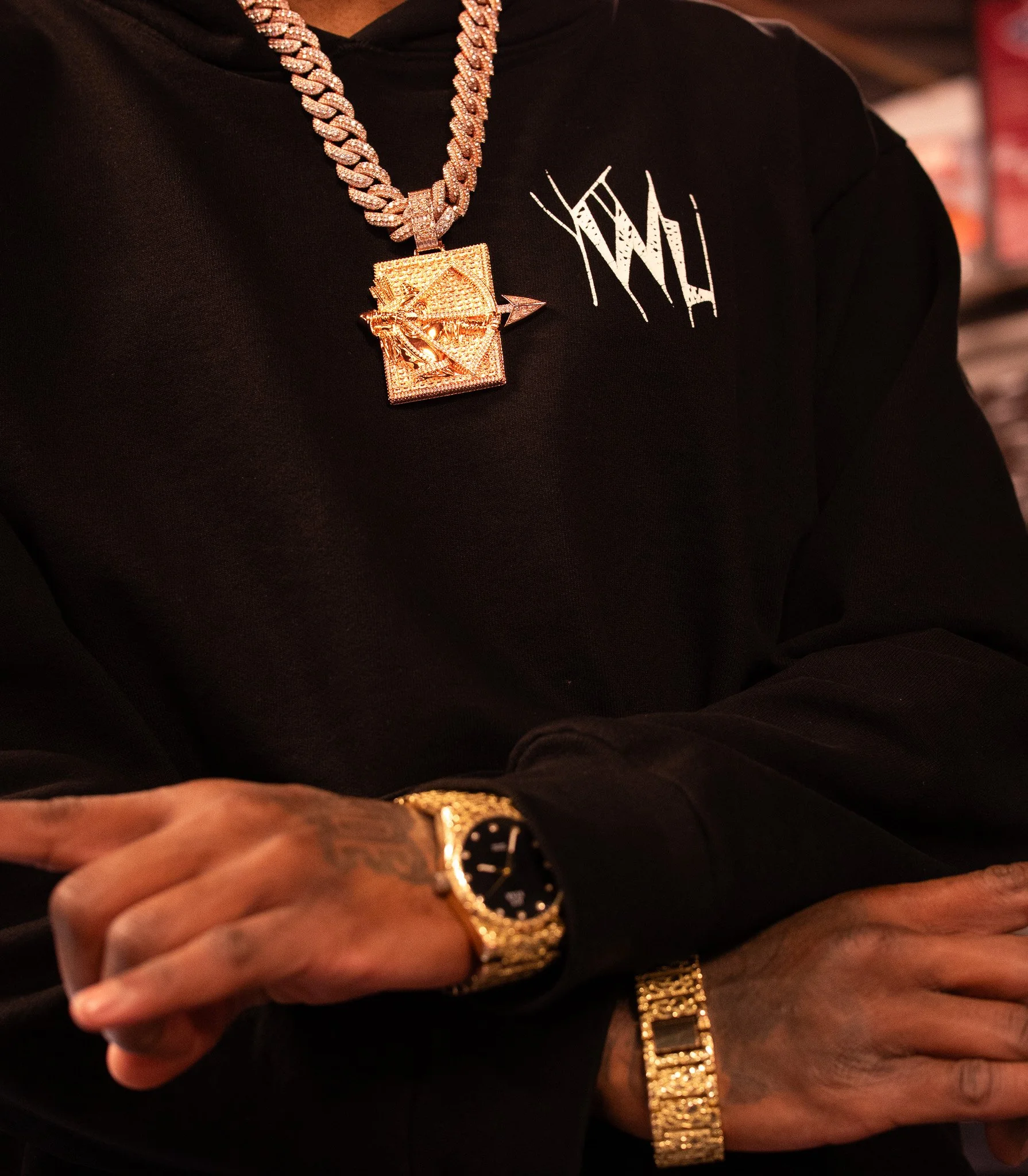 Close-up of a person wearing a black hoodie with white graphic text, adorned with a large gold chain necklace with a square pendant, a gold watch, and a gold bracelet. The person's hand is visible, showing tattoos.