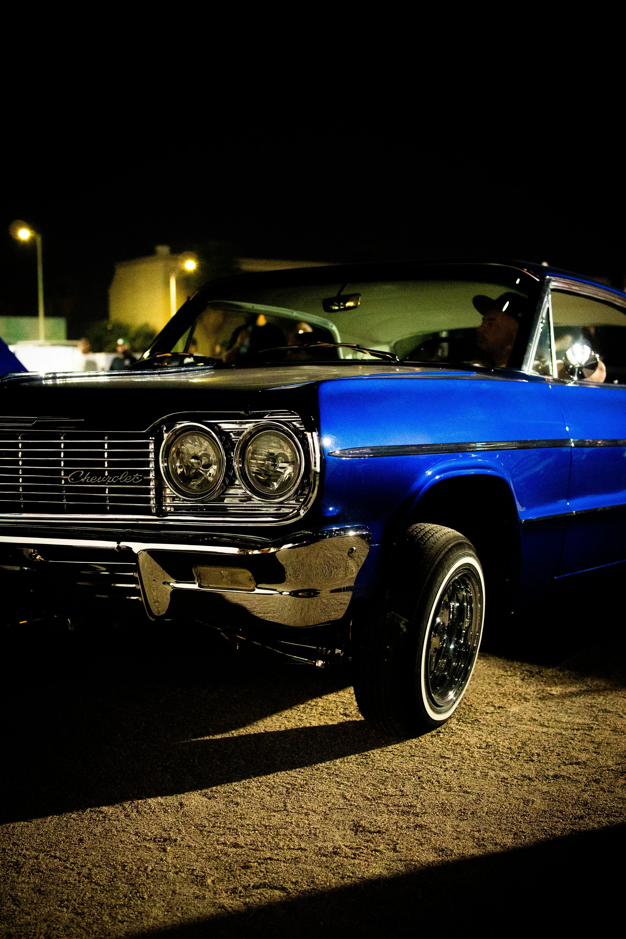 A classic blue Chevrolet car parked outdoors at night, with visible headlights, grille, and chrome bumper, and some people in the background.