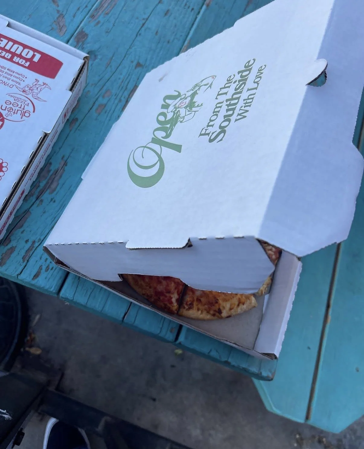 A white Pizza Hut pizza box with the lid partially open, revealing a pepperoni pizza inside, placed on a weathered blue wooden table.