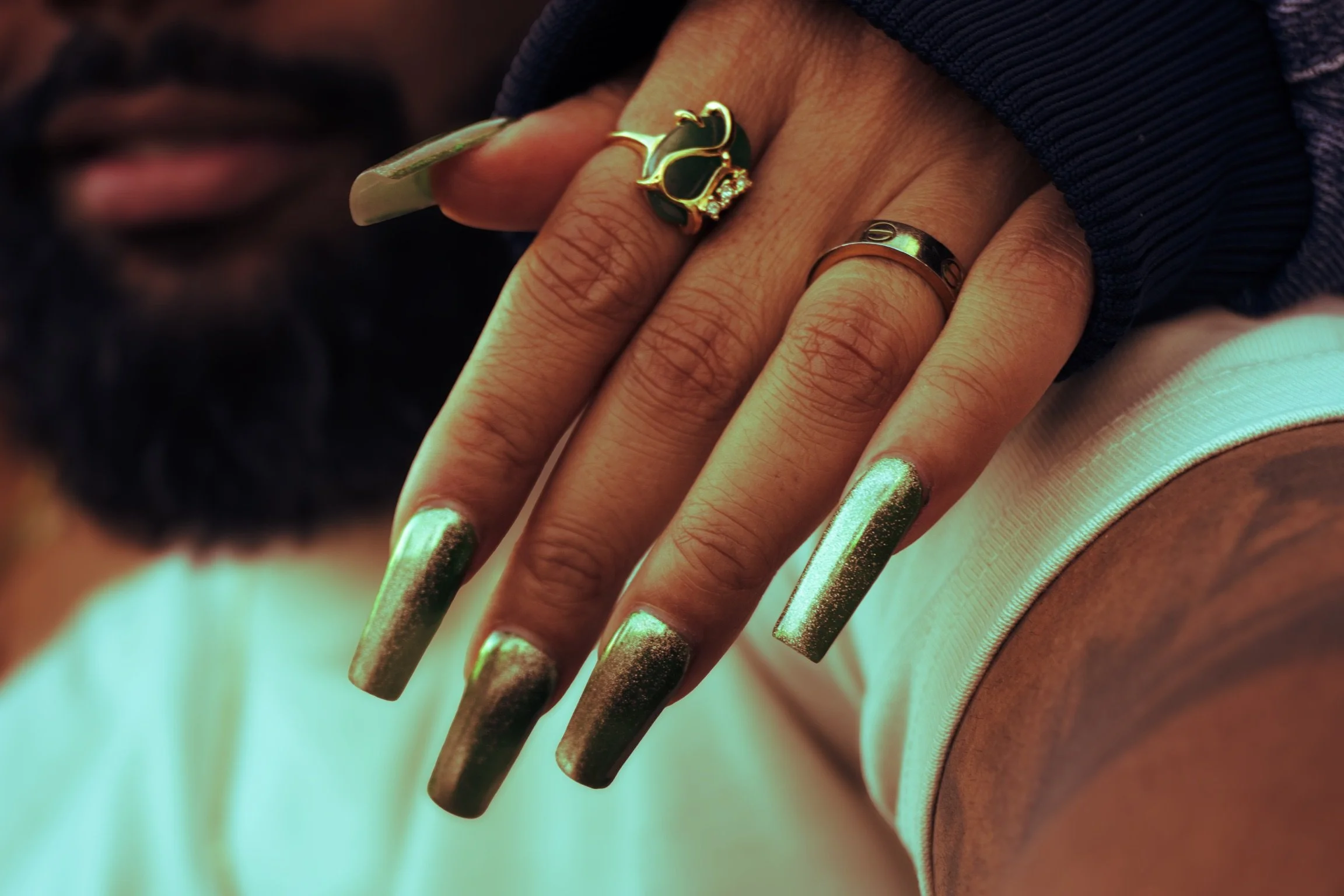 Close-up of a hand with long, metallic gold and black acrylic nails, featuring rings including a gold elephant ring and a black band, resting on a person's shoulder with a dark beard and lips visible in the background.