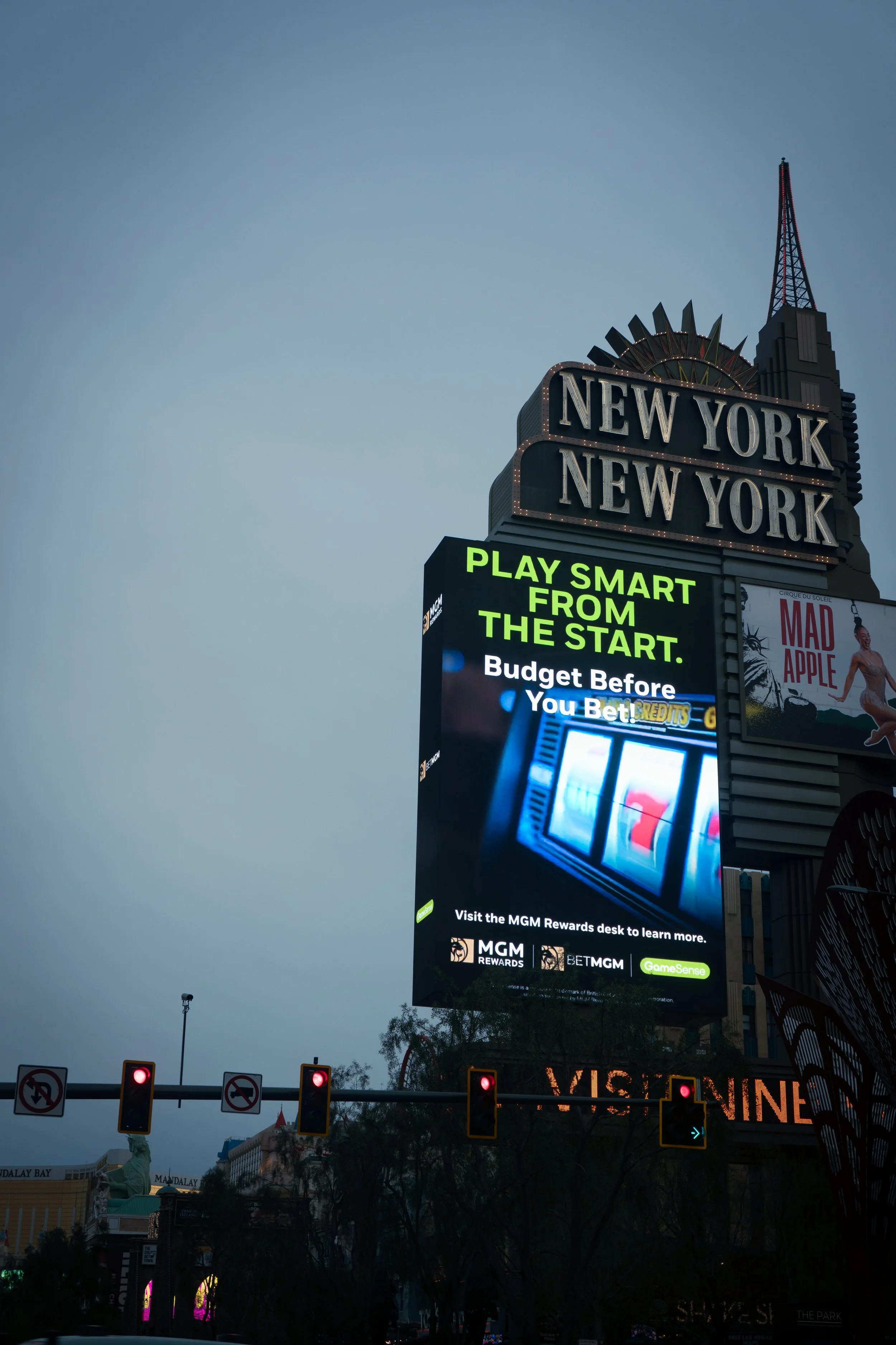 Bright electronic billboard advertisement in Times Square, New York, promoting smart gaming from the start, and a casino game with visible slot reels. The billboard is mounted above several traffic lights and street signs.