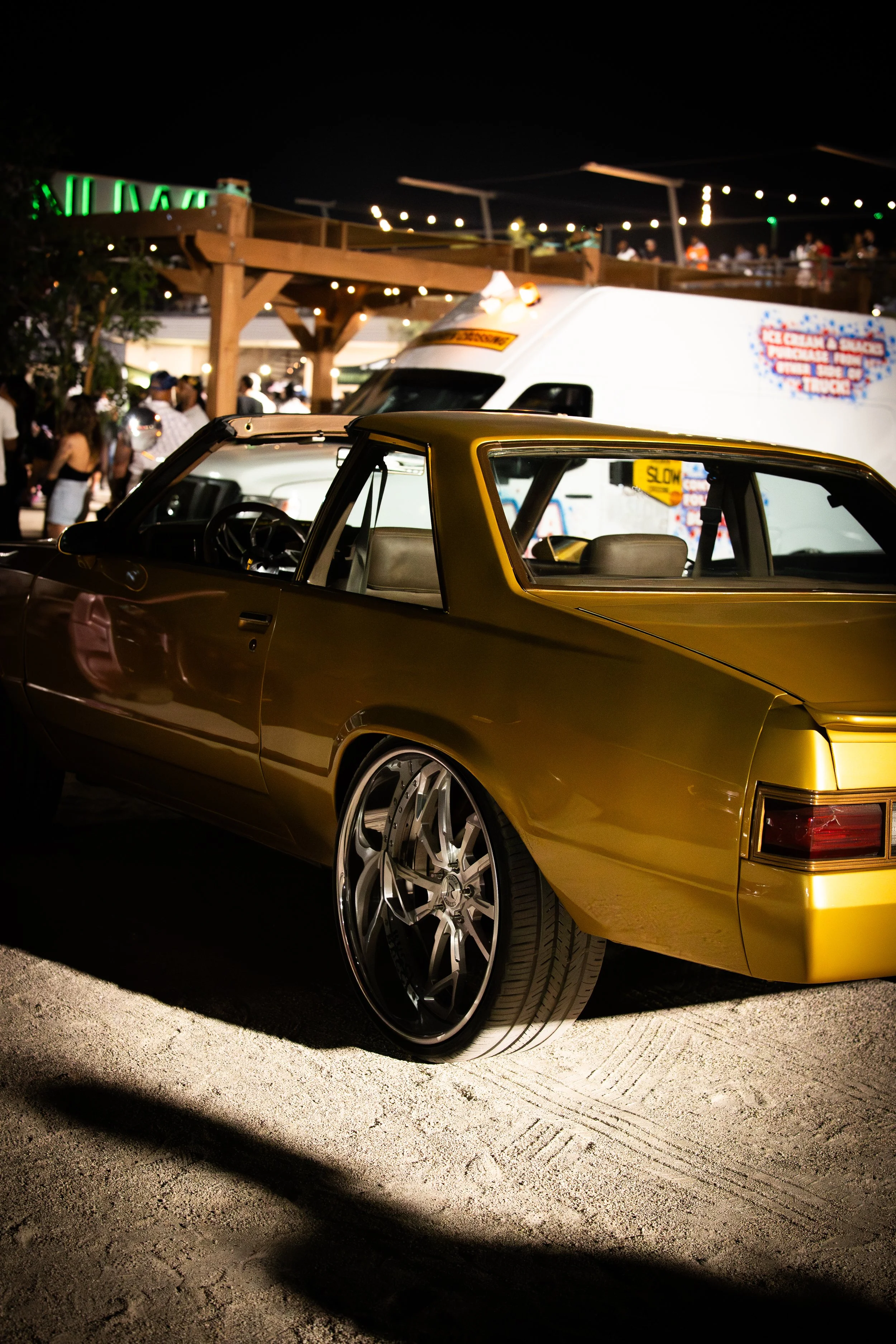 A gold-colored vintage car with custom large black rims at a nighttime outdoor event, with a stand and crowd in the background.