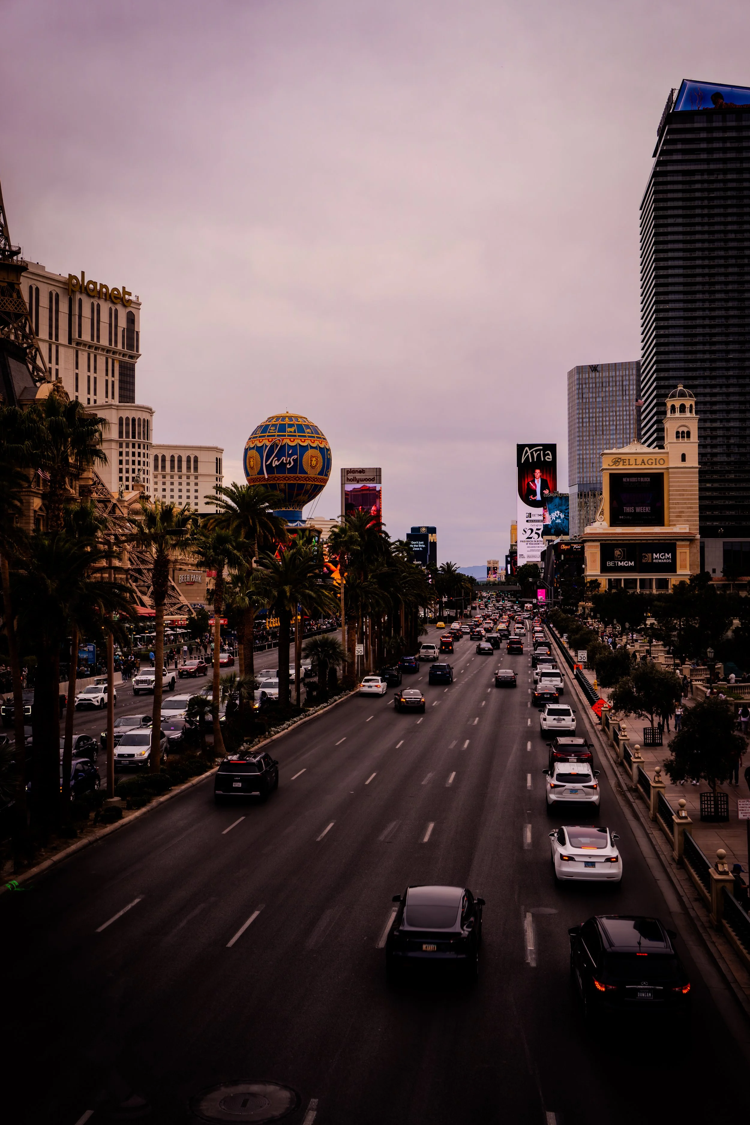 Downtown Las Vegas street scene at dusk with numerous cars on the road, palm trees lining the sidewalk, and buildings with neon signs, including the Paris Las Vegas hotel with its Eiffel Tower replica and a hot air balloon sign.