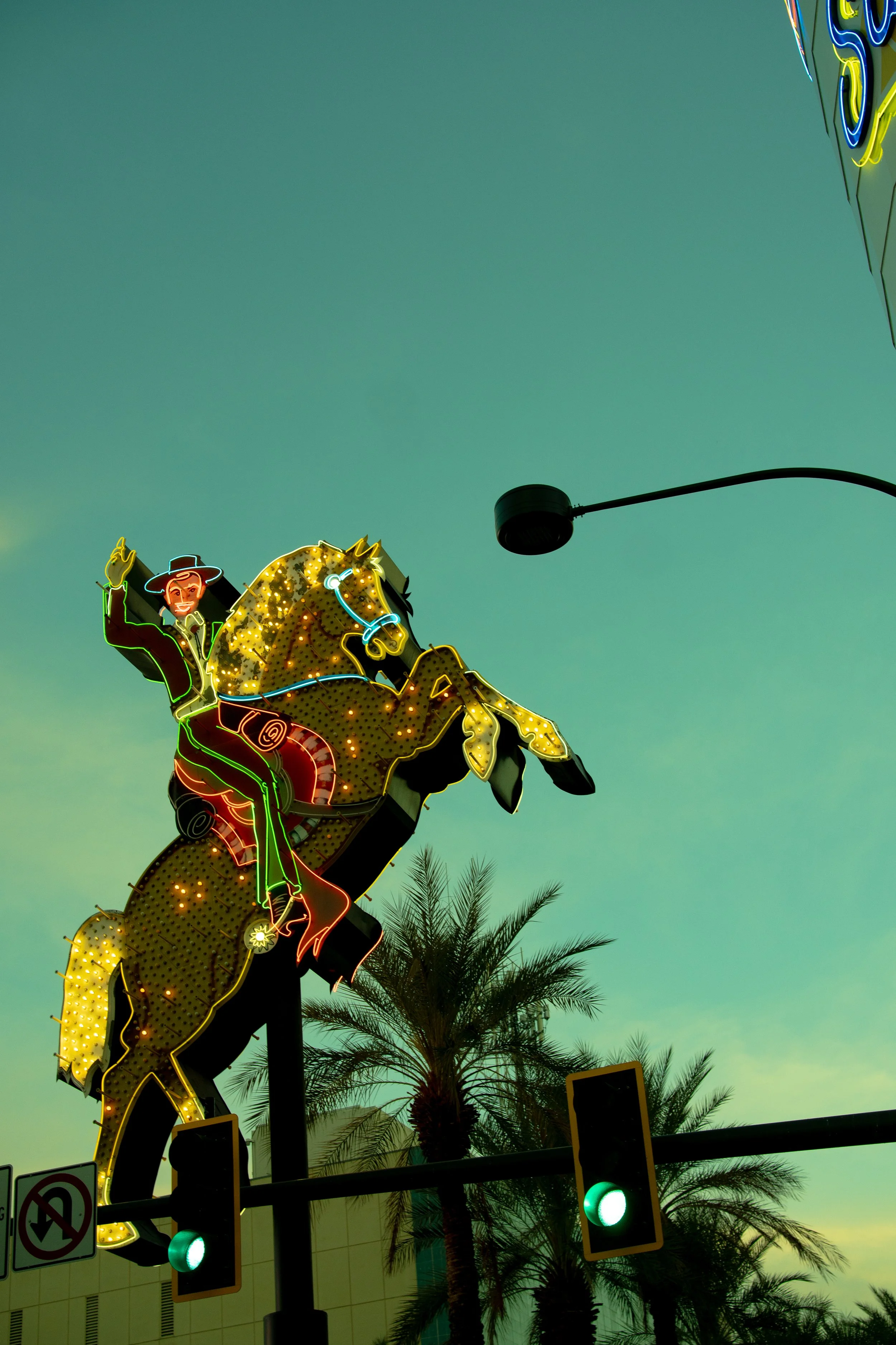 Colorful neon cowboy riding a yellow spotted horse, with palm trees and traffic lights in the background.