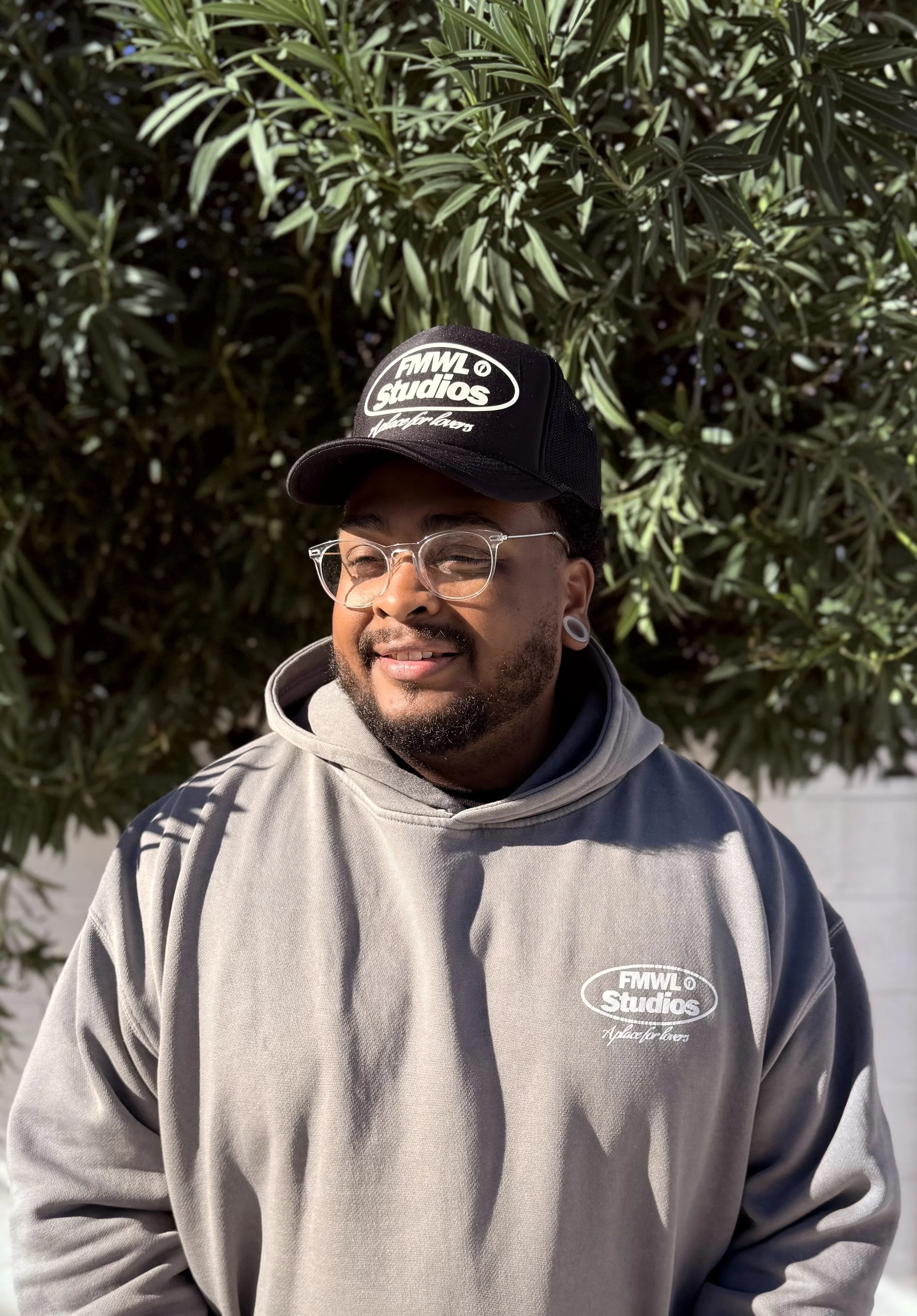 A man wearing glasses, a gray hoodie, and a black hat with 'FMWL Studios' logo stands outdoors in front of green leafy tree, smiling.