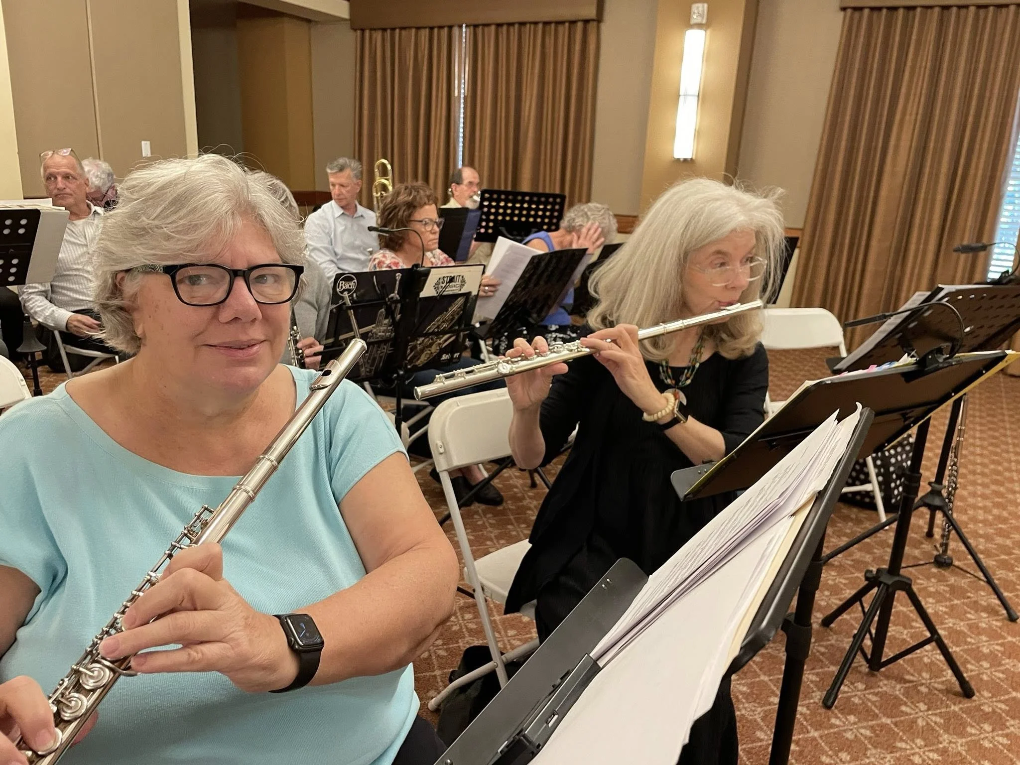 Two women, members of the New Horizons Band of Austin, Texas playing flutes during a concert warmup, with other musicians and music stands in the background.