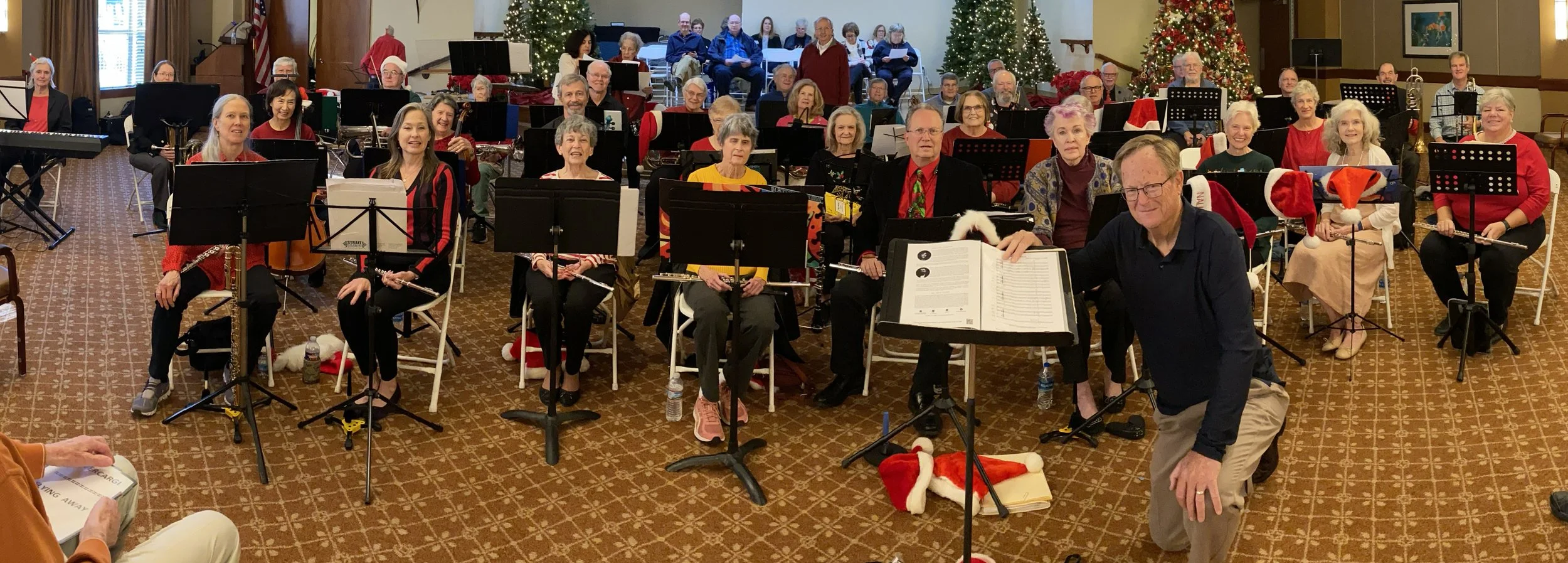 A holiday concert featuring a the New Horizons Band of Austin, Texas, some wearing Santa hats, with Christmas trees and festive decorations in the background