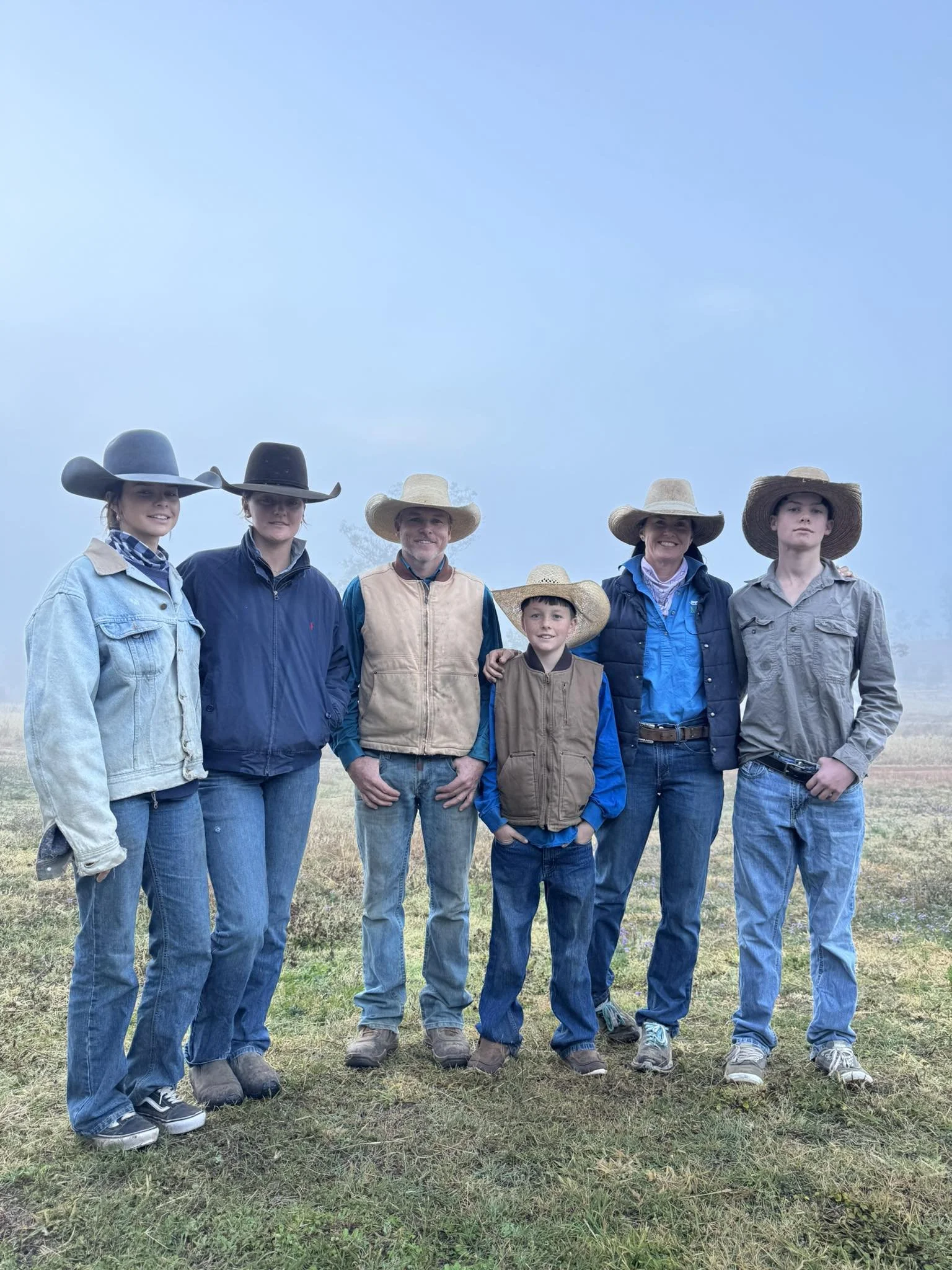A group of six people, three women, two children, and one man, standing outdoors in a foggy field, all wearing cowboy hats and casual clothing.