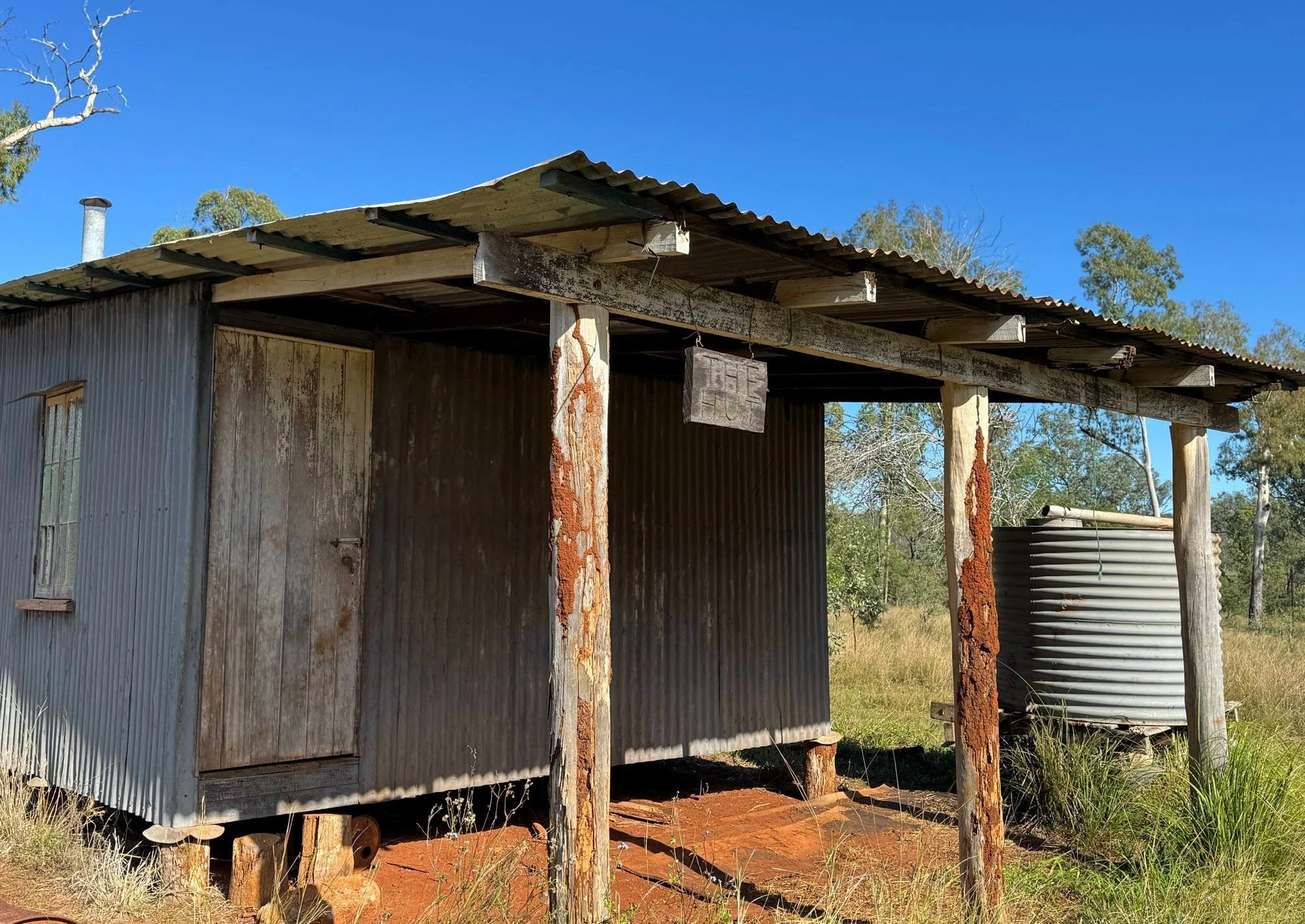 Keeping history intact. An old, weathered workers hut, handmande tin roof, wood shed with rusted support beams and corrugated metal roof in a rural area, with trees and grass in the background.