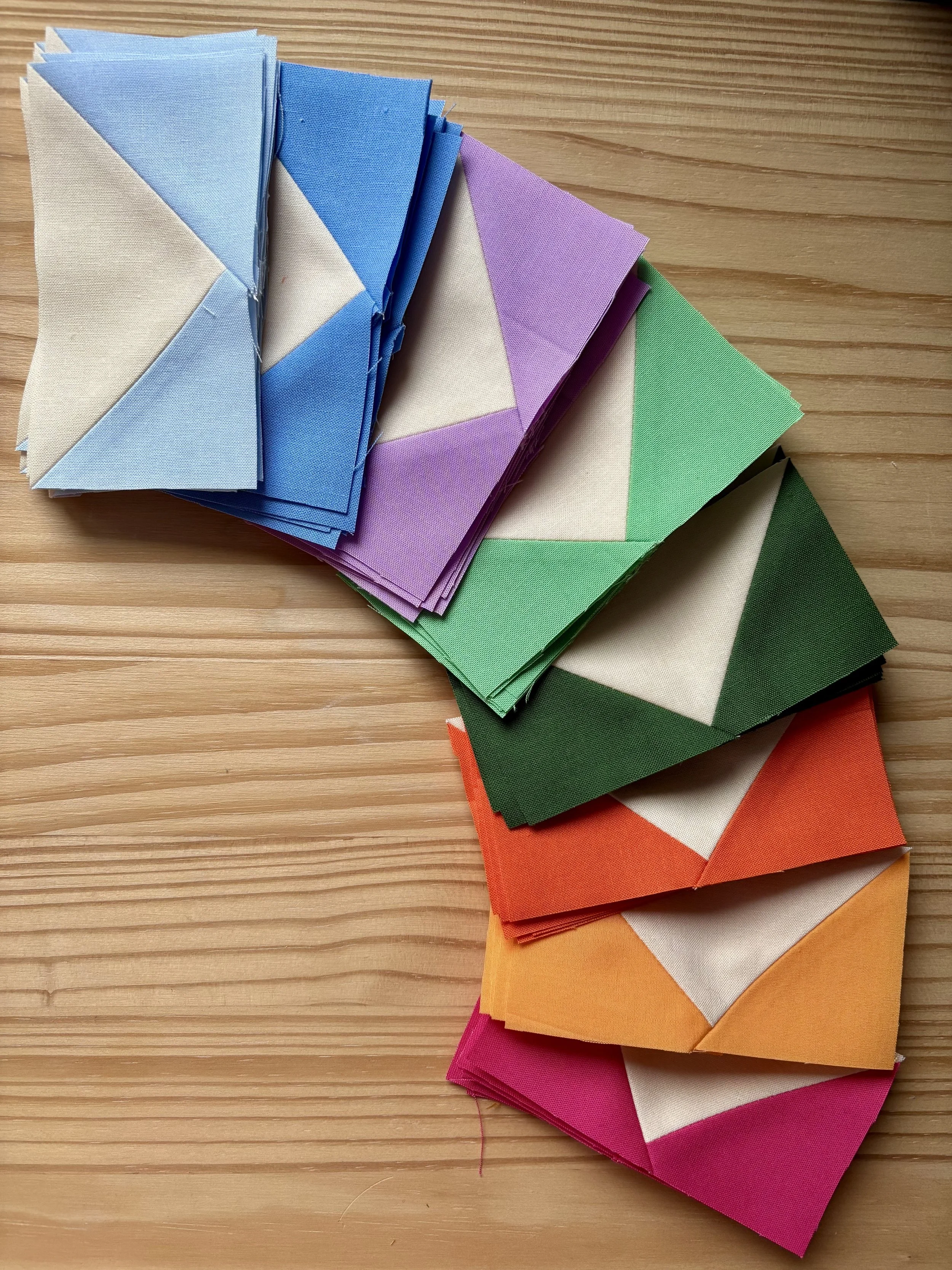 A  bird's eye view of a rainbow colour gradient of flying geese units with a cream background is fanned out on a natural pin wood surface.