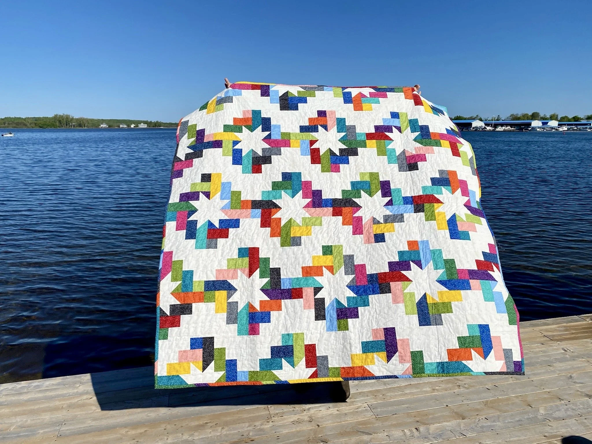 A vibrant colour completed quilt with 16 colours forming sawtooth stars is shown with a background view of a freshwater lake.