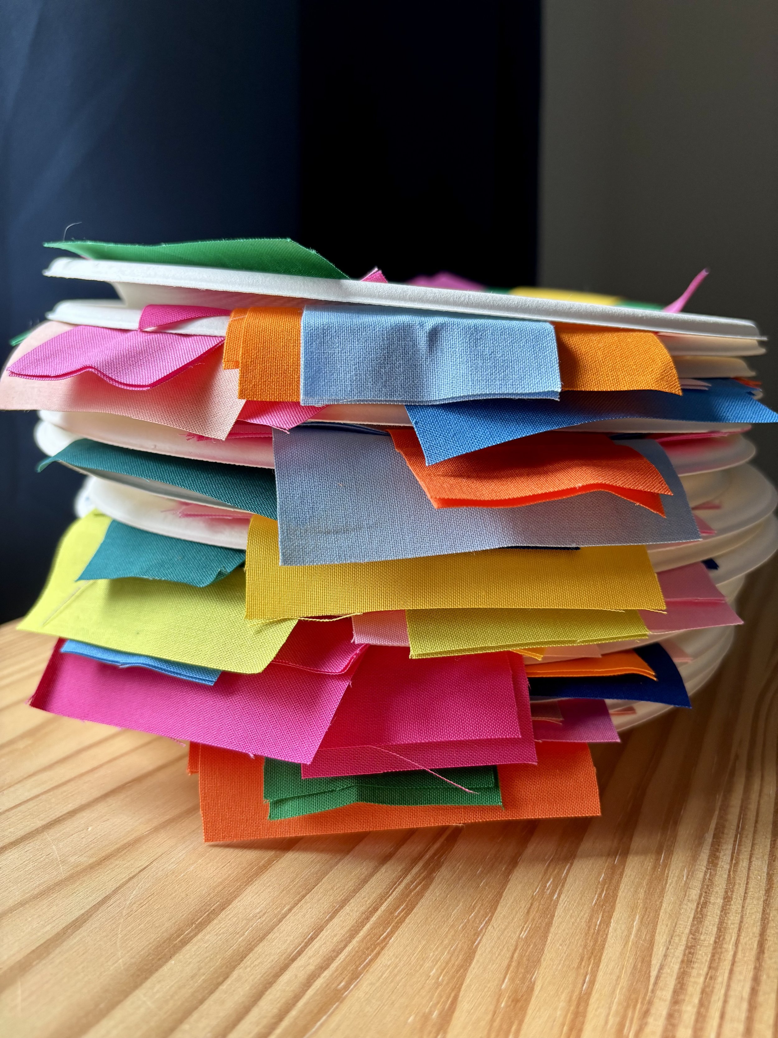 A stack of paper plates holding pieces of fabric that are hanging off the sides (hues of pink, blue, yellow, green, and orange)